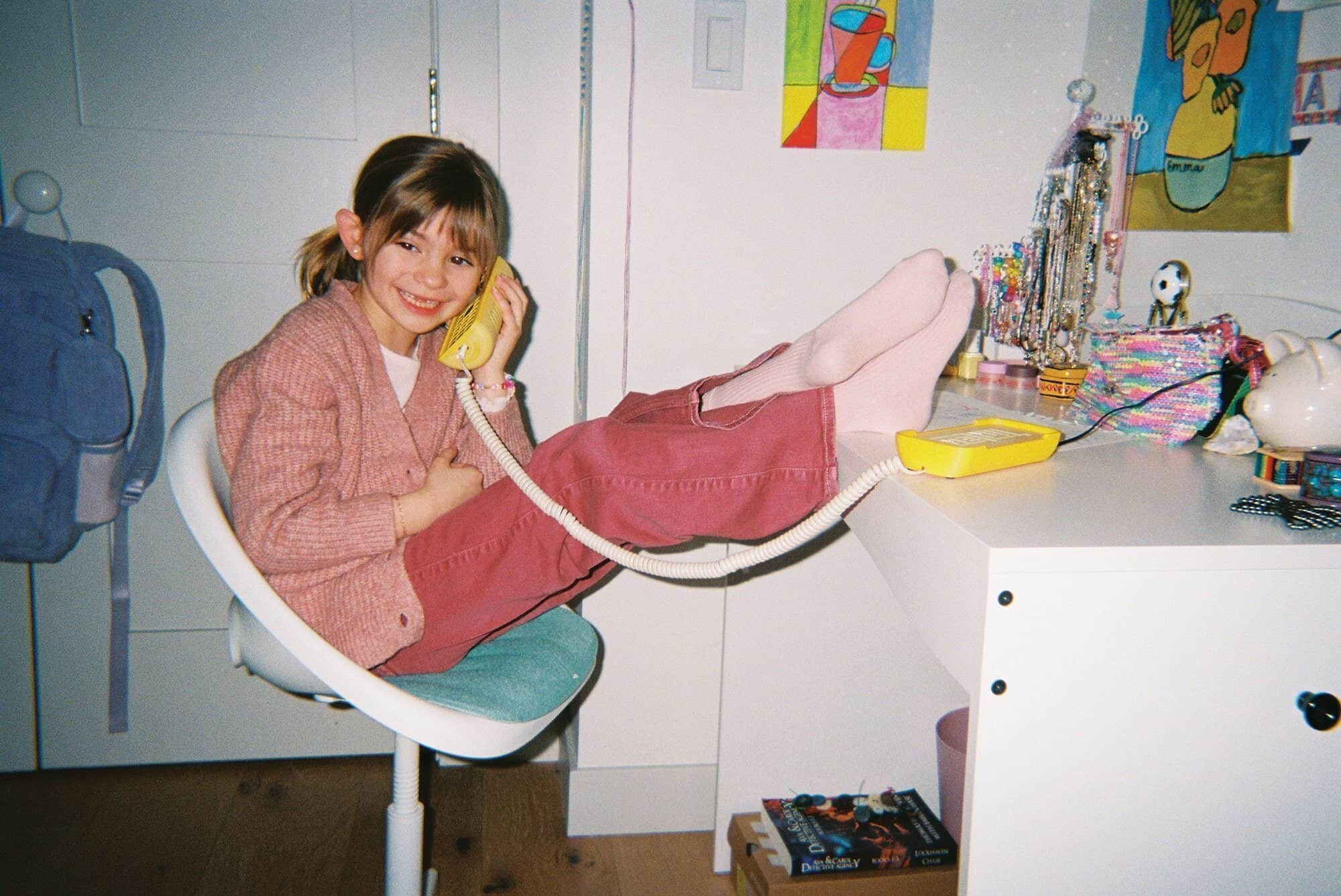 Young girl in pink clothes sits with feet up on a desk, smiling and talking on a yellow corded phone in a colorful bedroom, radiating joy like the spirit of "Merry Christmas Baby.
