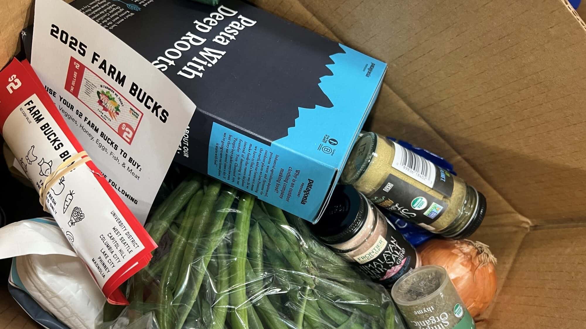 A cardboard box containing green beans, a yellow onion, spice jars, a cookbook, and a 2025 Farm Bucks flyer.