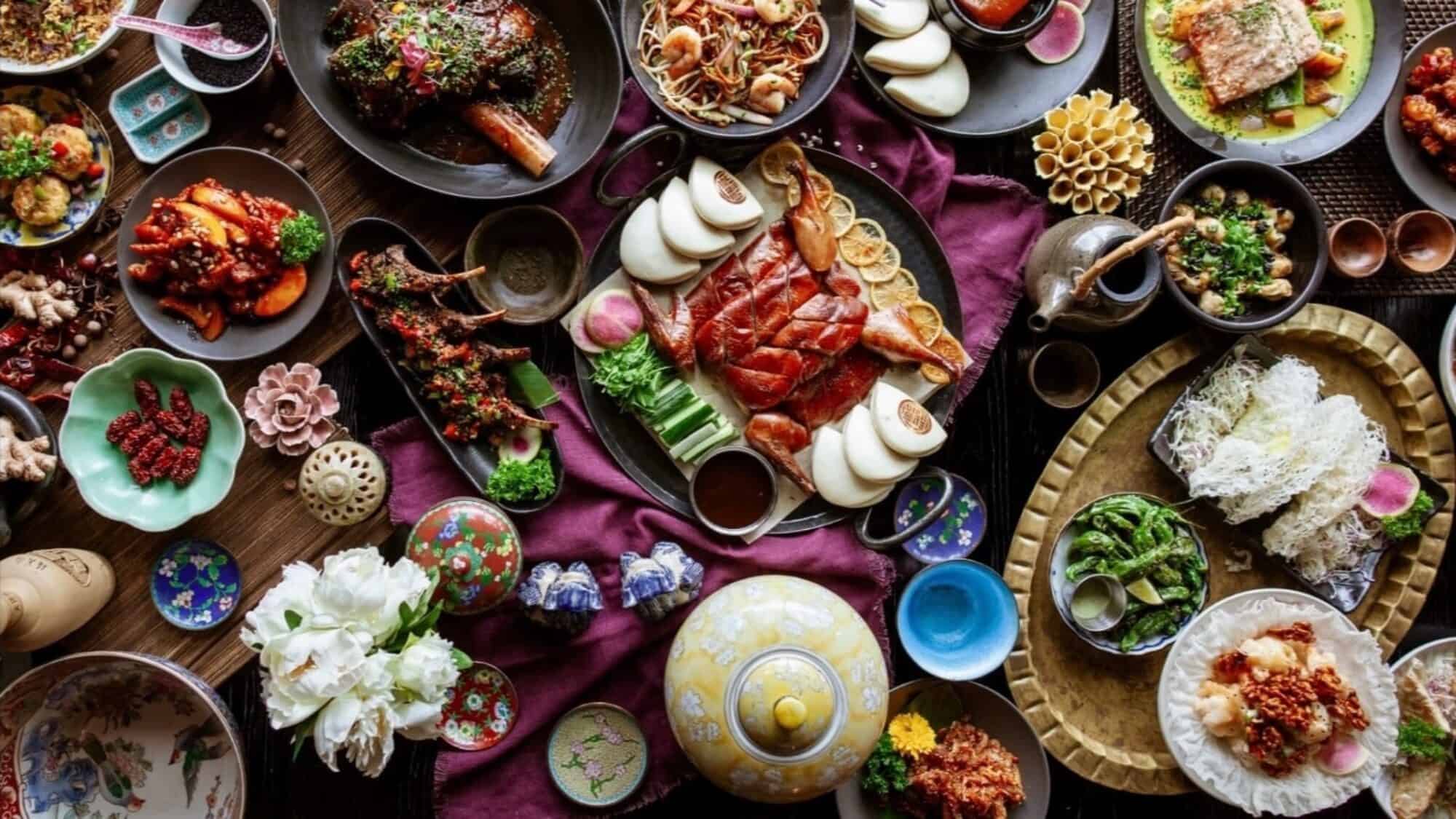 A variety of Chinese dishes, including roast duck, steamed buns, seafood, vegetables, and sauces, are arranged on a table with decorative plates and teapots—perfect for a unique Thanksgiving dining experience.