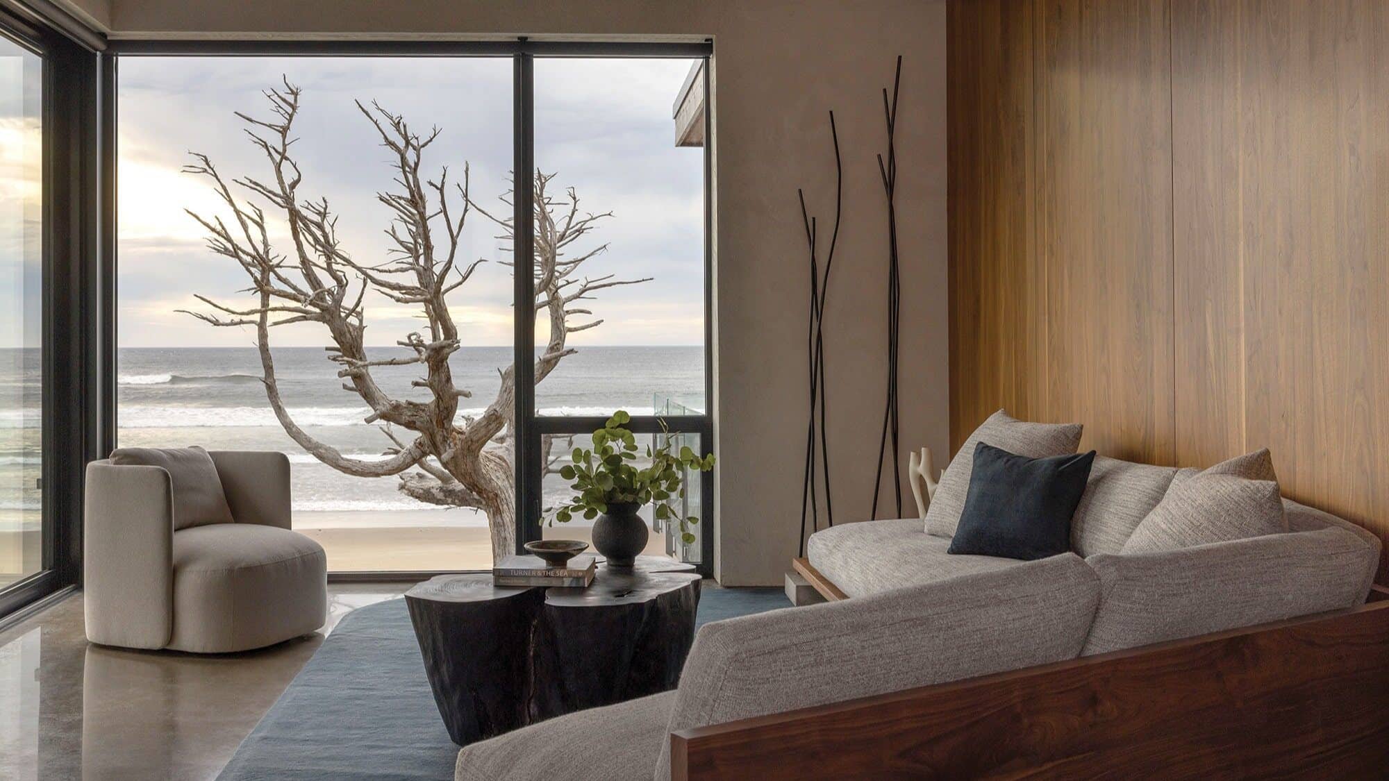 Modern living room with grey sofa, wood accents, and a coffee table overlooks the ocean near Arch Cape through large glass doors. A leafless tree is visible on the balcony outside, evoking a cozy Oregon beach retreat.