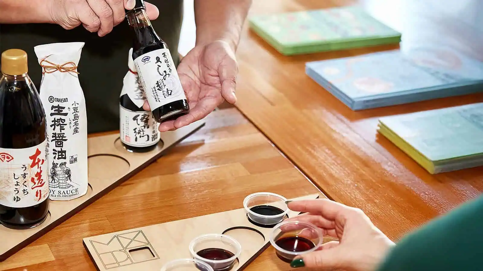 People sample different soy sauces in small cups, with various bottles and packages of Japanese soy sauce displayed on a wooden table.