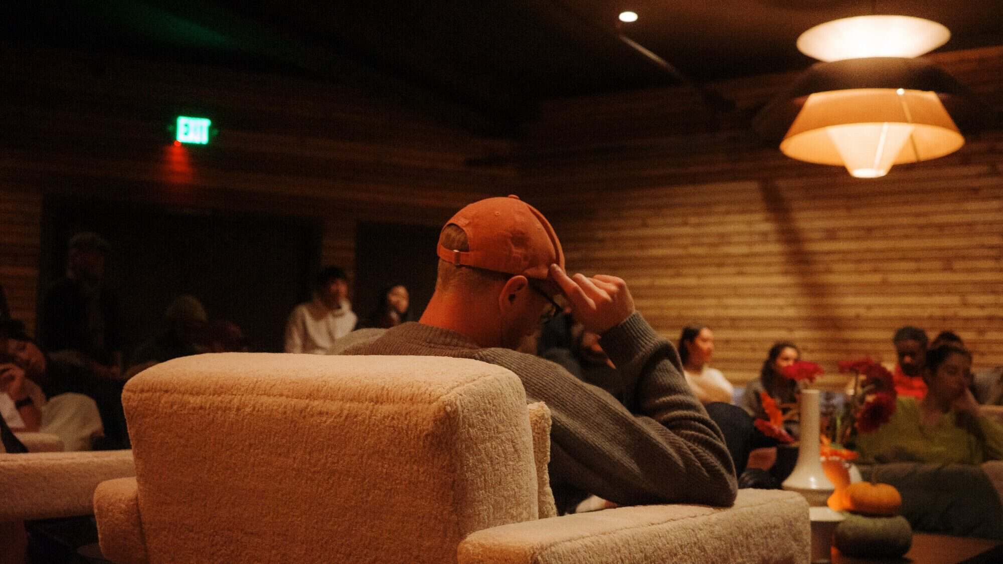 A person in an orange cap sits in a beige armchair facing a group of people in a dimly lit room with wooden walls and modern lighting.