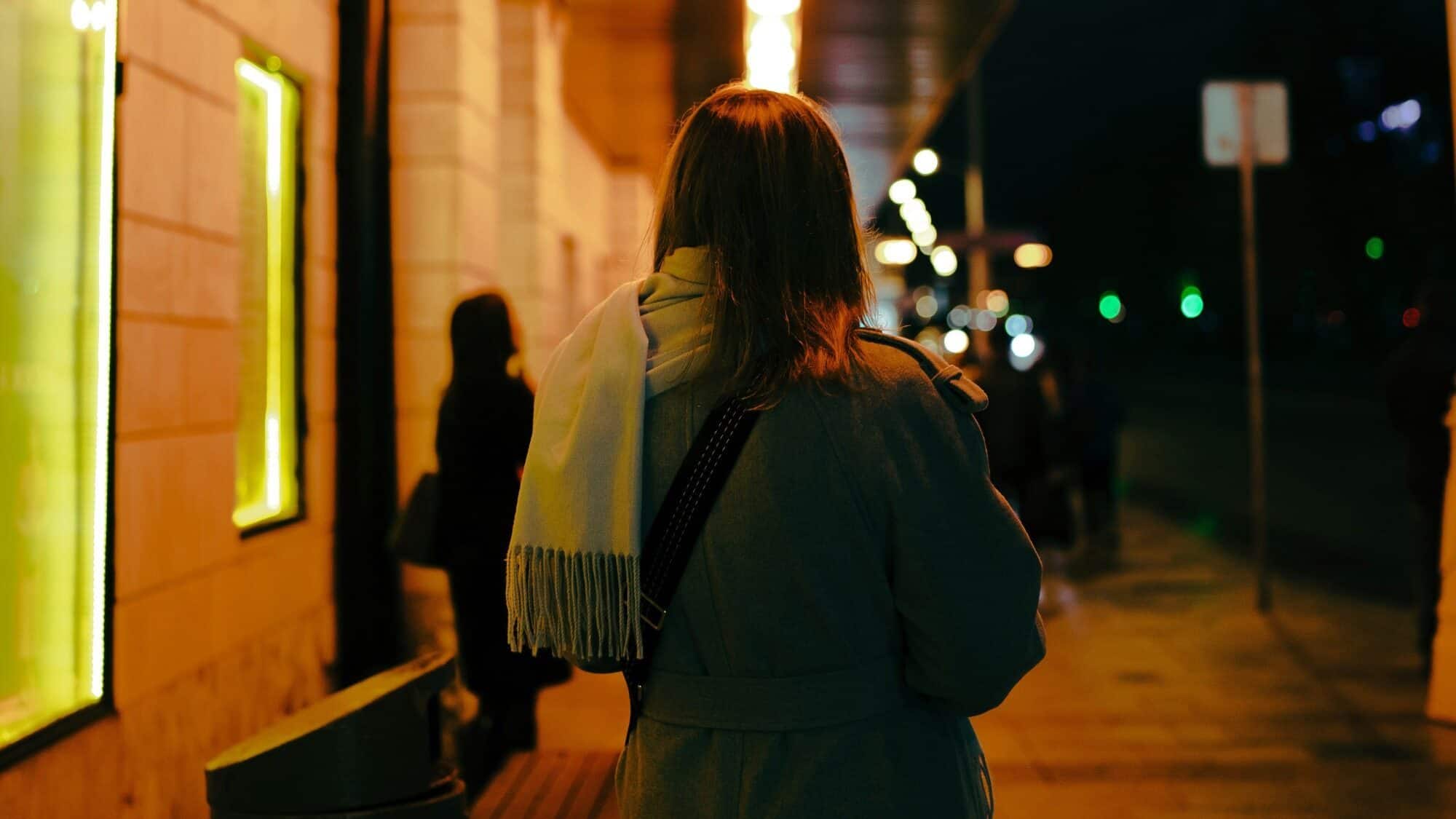 A person wearing a coat and scarf walks alone on a city sidewalk at night; lit shop windows and streetlights line the street.