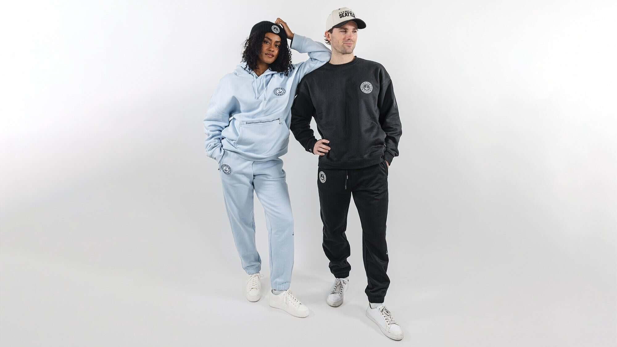 Two people stand against a white background wearing matching Little Escapes sweatsets and white sneakers; one in light blue and the other in black, both featuring logos and baseball caps from our Fave Five collection.