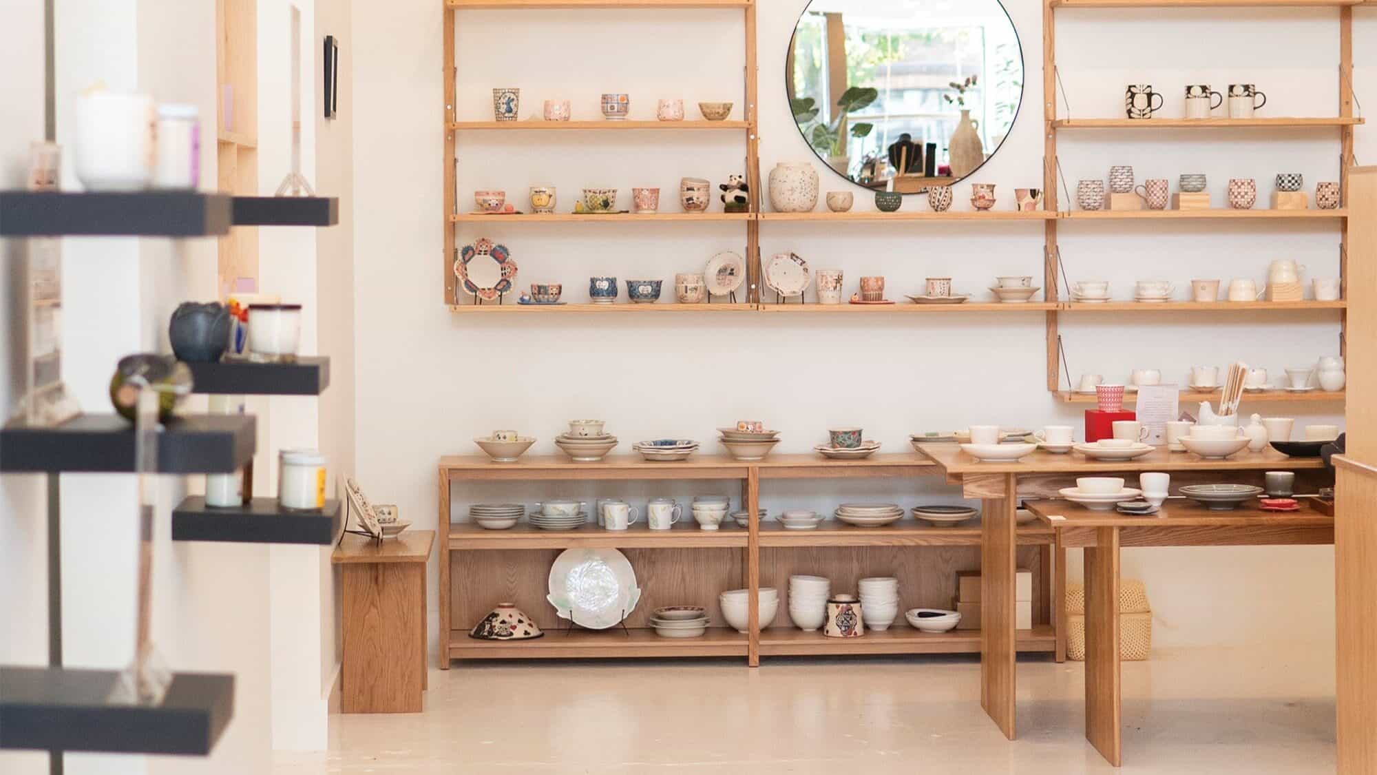 A ceramics shop interior from Little Escapes features wooden shelves and tables displaying bowls, cups, and plates, with a round mirror on the wall reflecting greenery from outside.