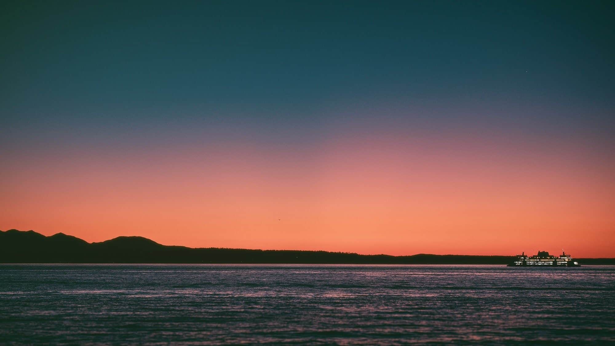 A ferry crosses calm water at sunset with mountains silhouetted in the background and a gradient sky from orange to deep blue, capturing a moment of tranquility perfect for photography enthusiasts like Nick Ward.