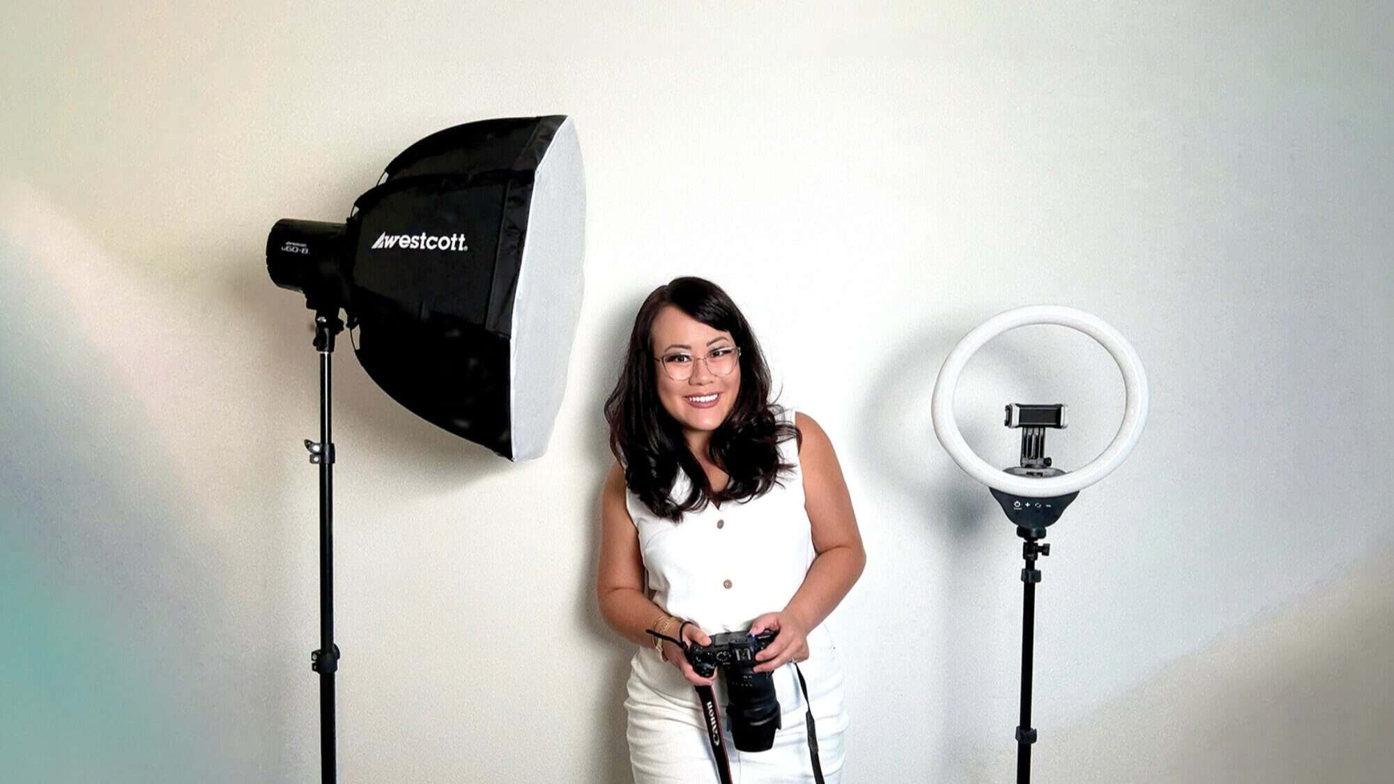A woman in white stands between a studio softbox light and a ring light, holding a camera with a sense of future thinking, in front of a plain white wall.