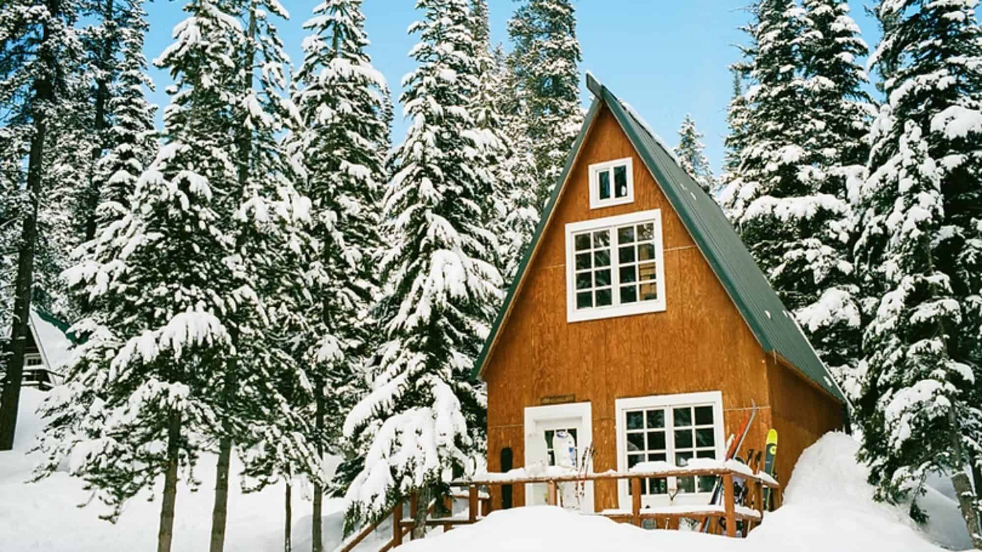 A wooden A-frame cabin surrounded by tall snow-covered pine trees under a clear blue sky. Snow blankets the ground and the cabin’s roof.