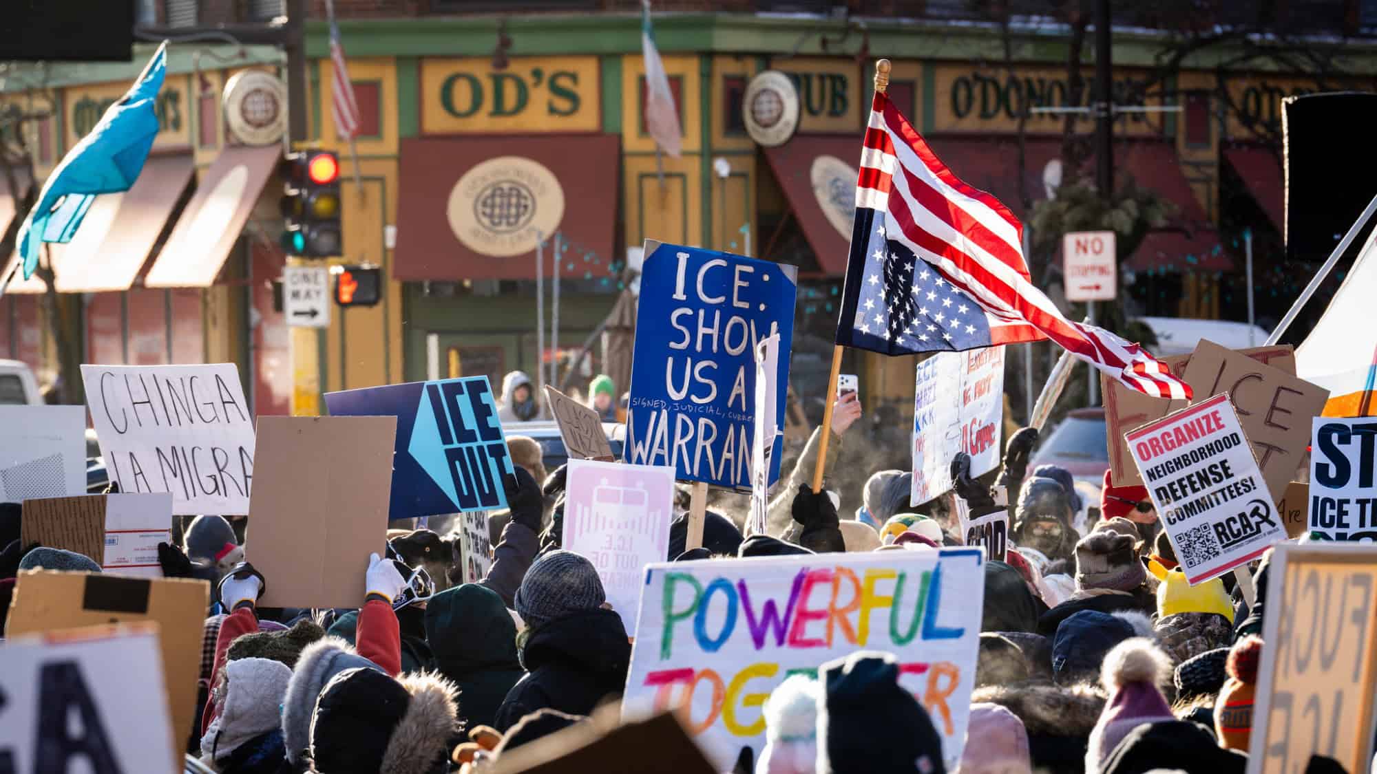 A crowd of protesters holds various signs, including messages to ICE and support for immigrants, in front of colorful Minneapolis storefronts. An American flag is visible among the signs during a National Shutdown demonstration.