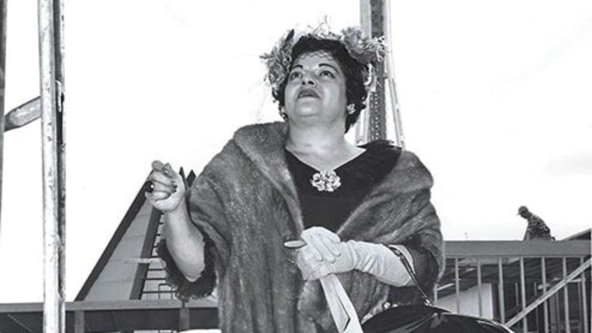 A woman in a fur shawl and gloves stands outdoors, holding a handbag and umbrella, with a building and staircase in the background.
