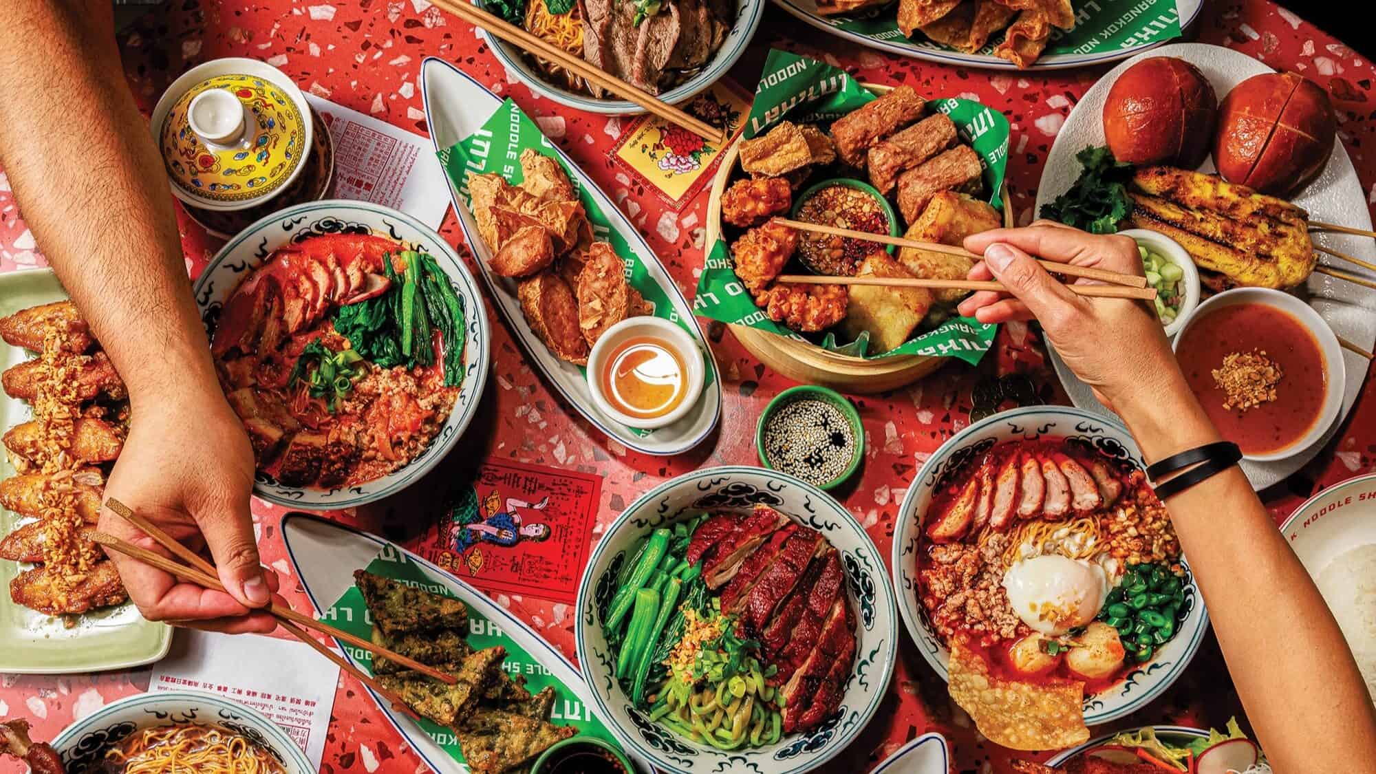 A colorful spread of assorted Asian dishes, including rice bowls, grilled meats, vegetables, and dipping sauces, with hands using chopsticks to serve food.