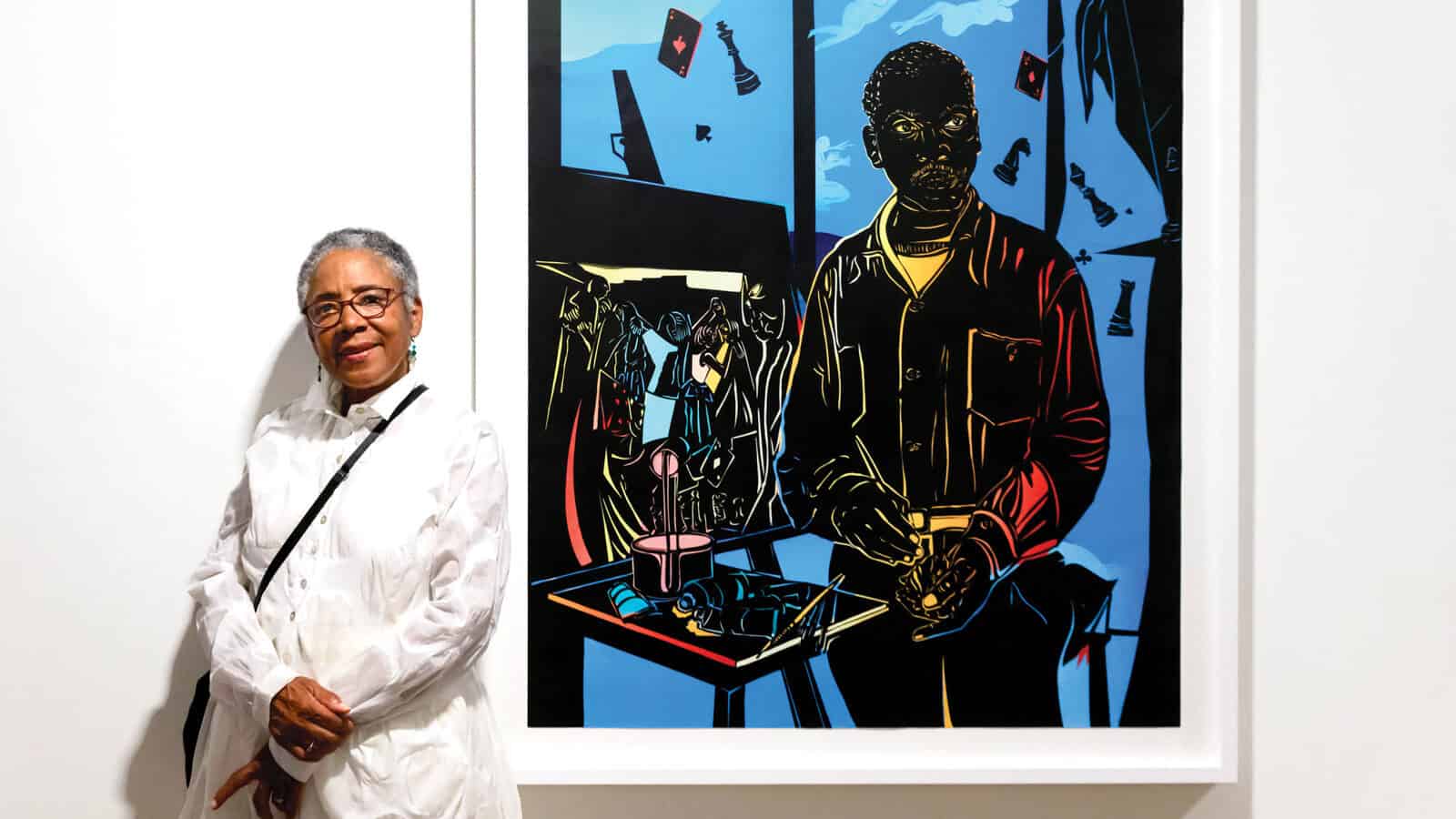 An older woman in a white outfit, Cultural Torchbearer Barbara Earl Thomas, stands next to a colorful framed painting of a person at a table, displayed on a gallery wall.