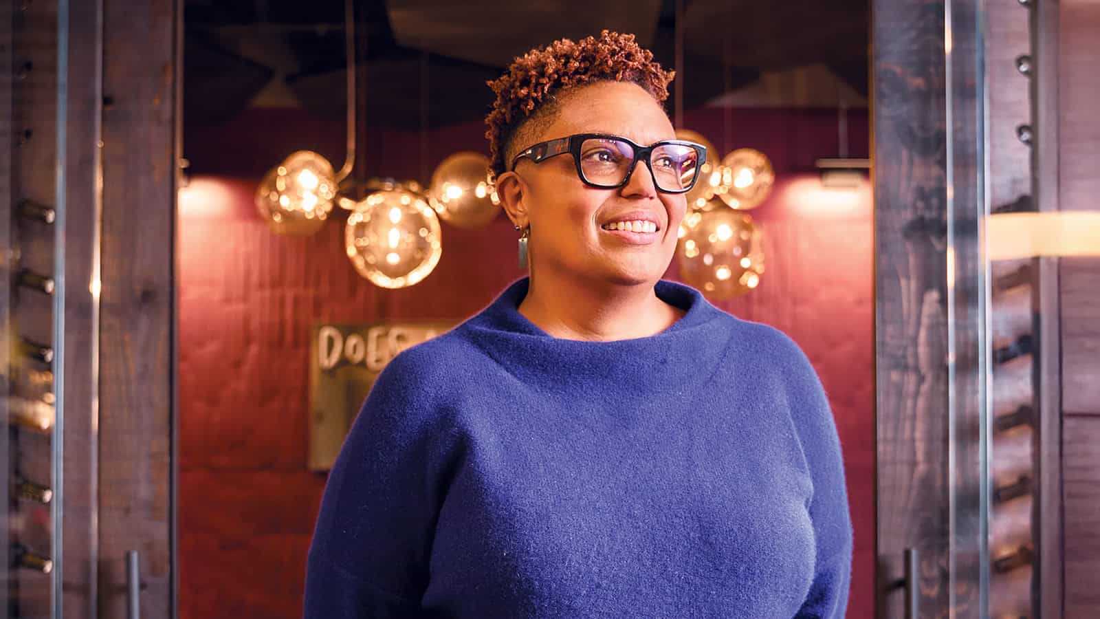 Jamila Conley, wearing glasses and a blue sweater, stands indoors smiling. Round light fixtures and an Industry Expander sign are blurred in the background.