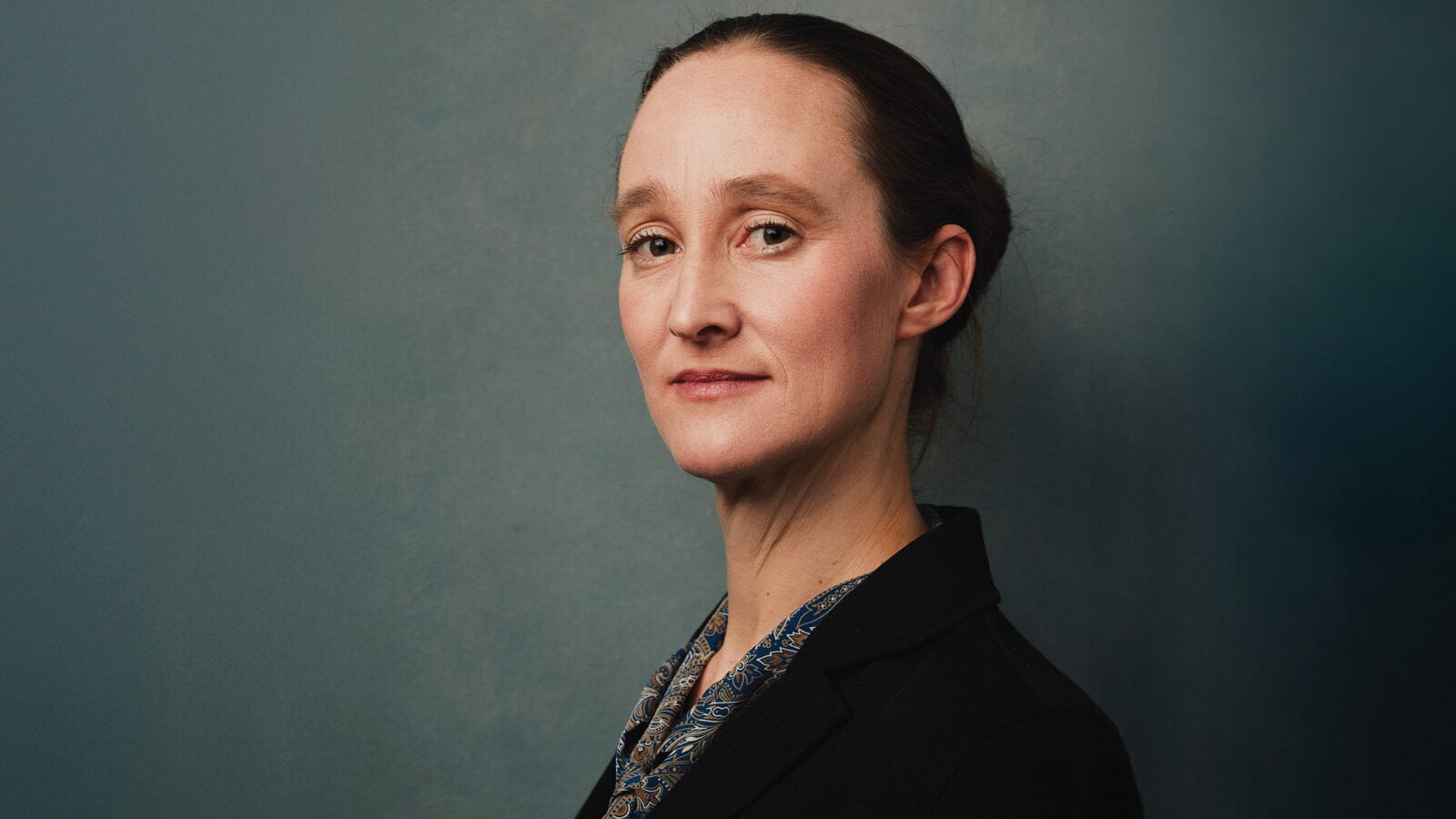 Katie Wilson, with her hair tied back, wears a dark blazer and patterned shirt as she stands against a plain blue-grey background, looking slightly to the side—an image befitting The Mayor.