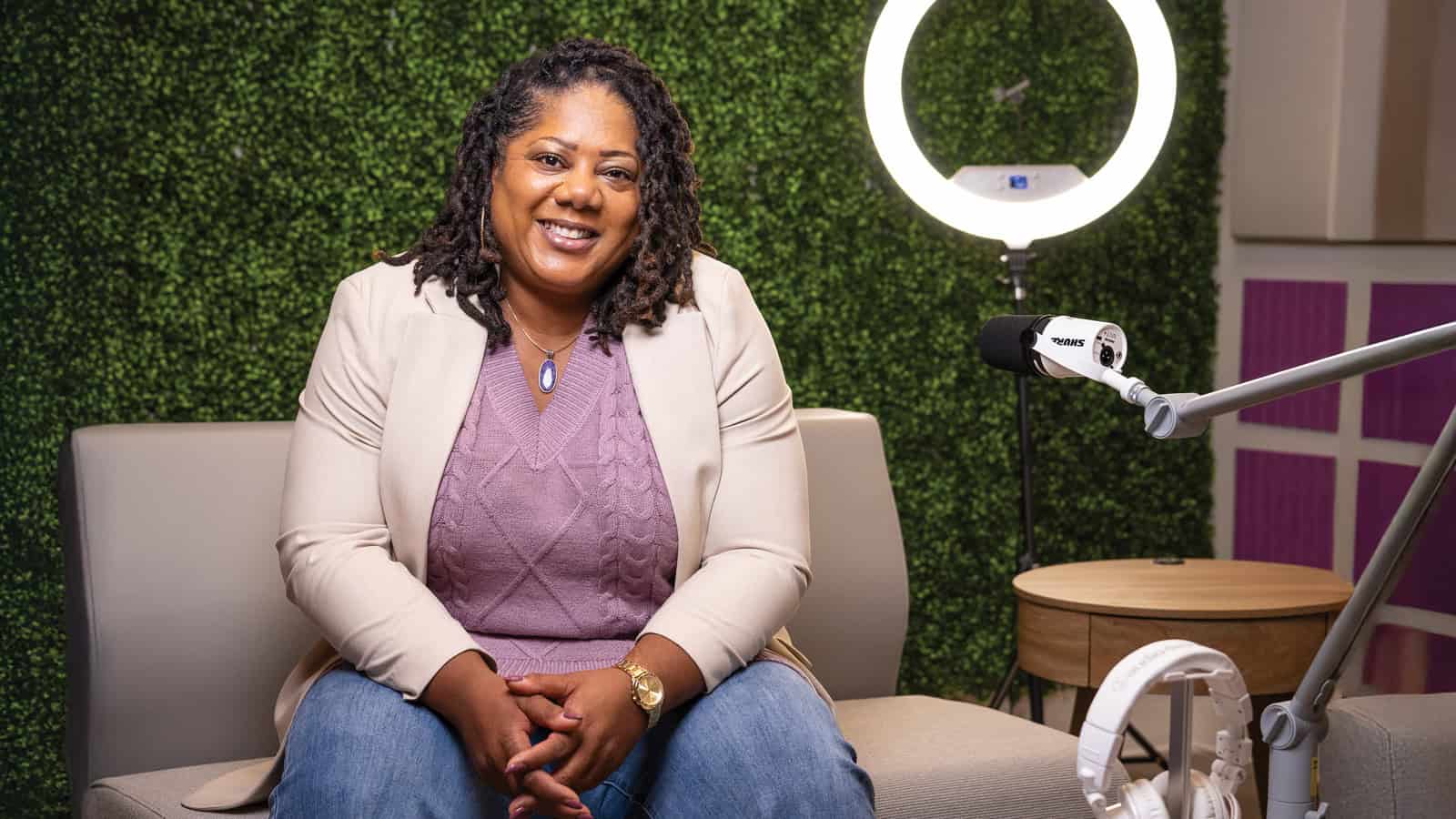 A woman sits on a couch in front of a green wall, smiling at the camera; a ring light and microphone are set up nearby as Nicky Gane of Dignity for Divas prepares to share her inspiring story.