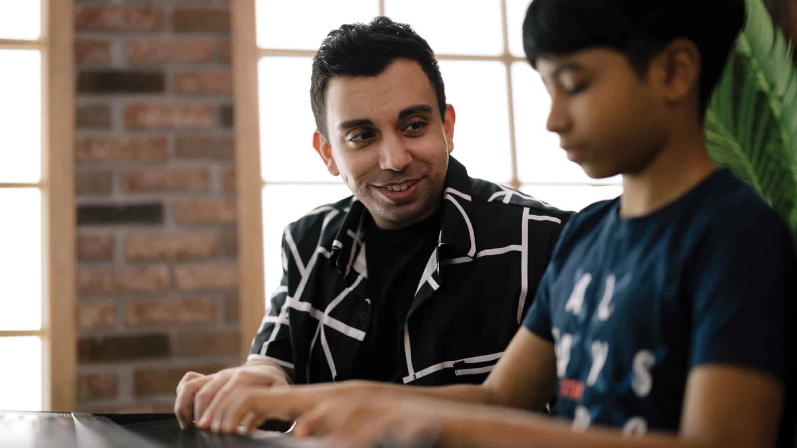 An adult, possibly a piano teacher like Payam Khastkhodaei, sits beside a child at a table. The adult smiles warmly as the child focuses intently on the laptop screen.