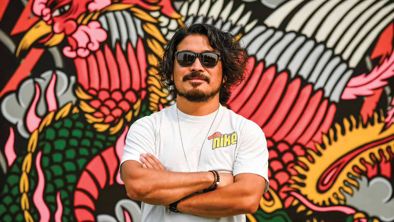 A man wearing sunglasses and a white Nike t-shirt stands with arms crossed in front of a colorful dragon mural by artist Shogo Ota.