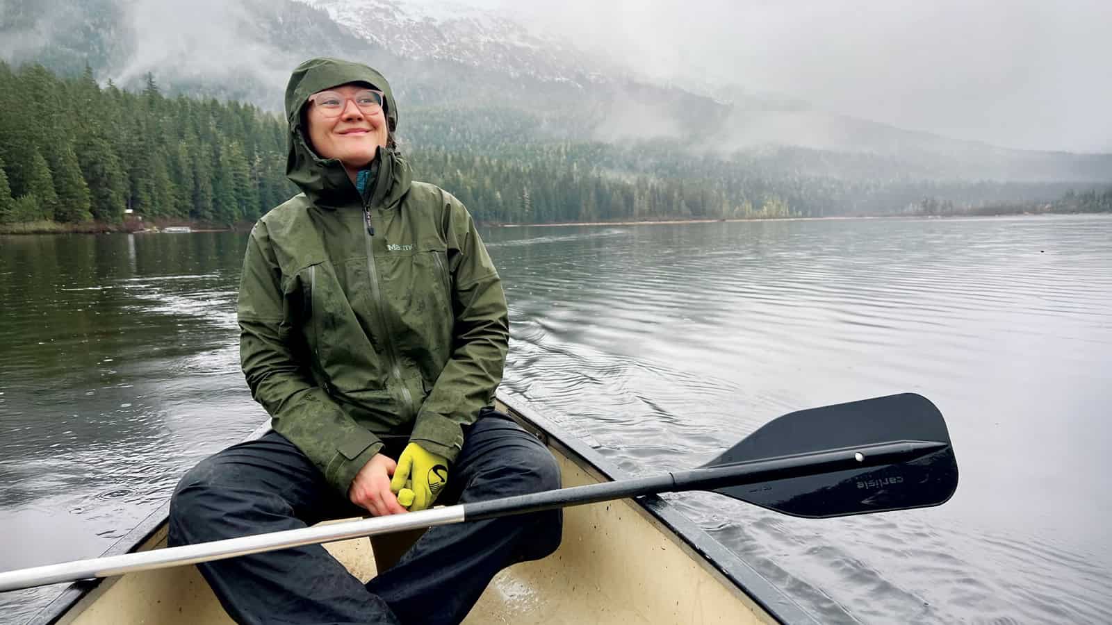 A person, embodying The Explorer, wears a green rain jacket and glasses while sitting in a canoe on a calm lake, surrounded by forest and misty mountains, holding a paddle. Inspired by adventurers like Tessa Hulls.
