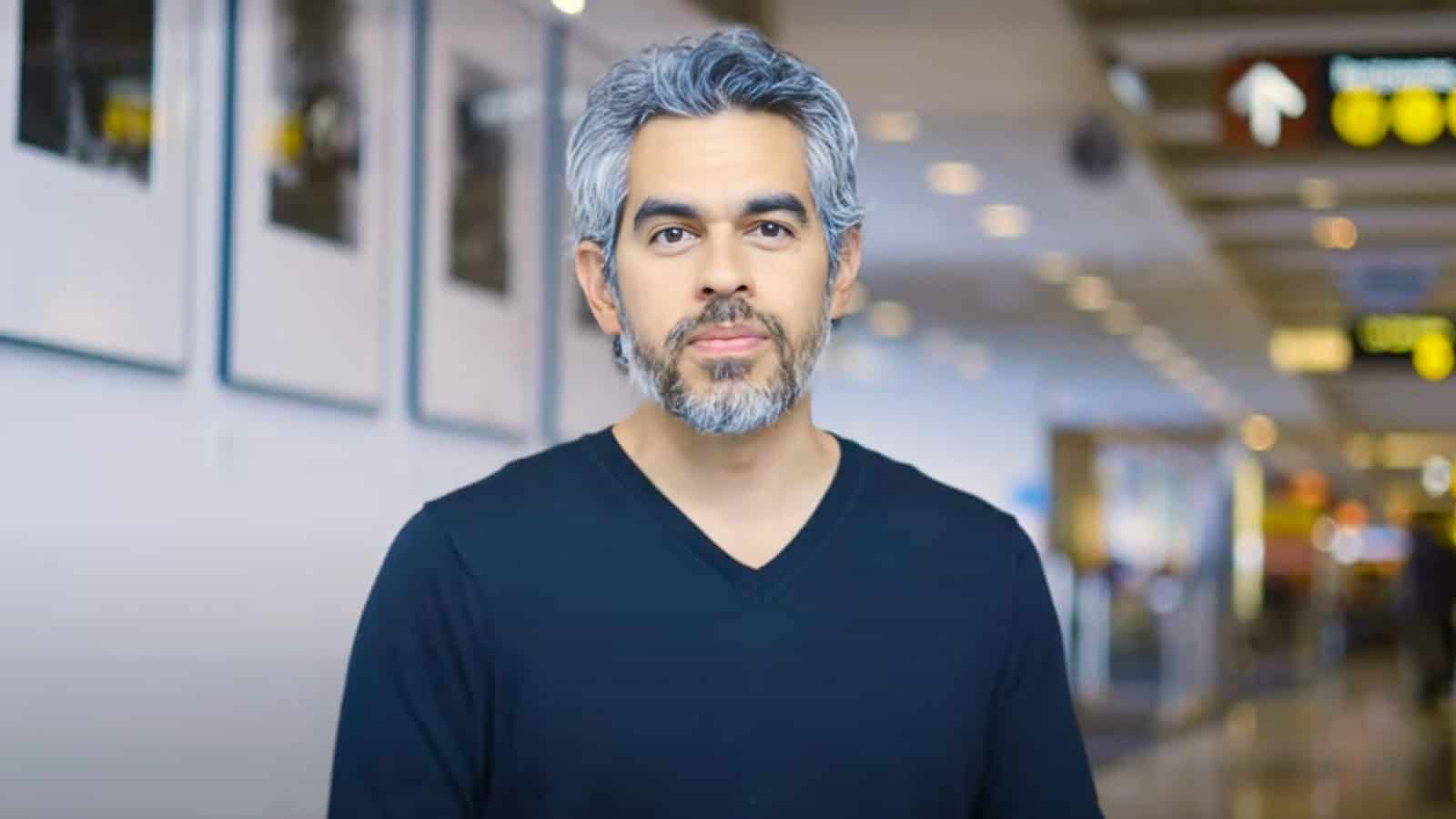 A man with gray hair and a beard, wearing a black V-neck shirt, stands indoors in a brightly lit hallway with signs and framed pictures—Civic Spacemaker Tommy Gregory is captured in this thoughtful moment.