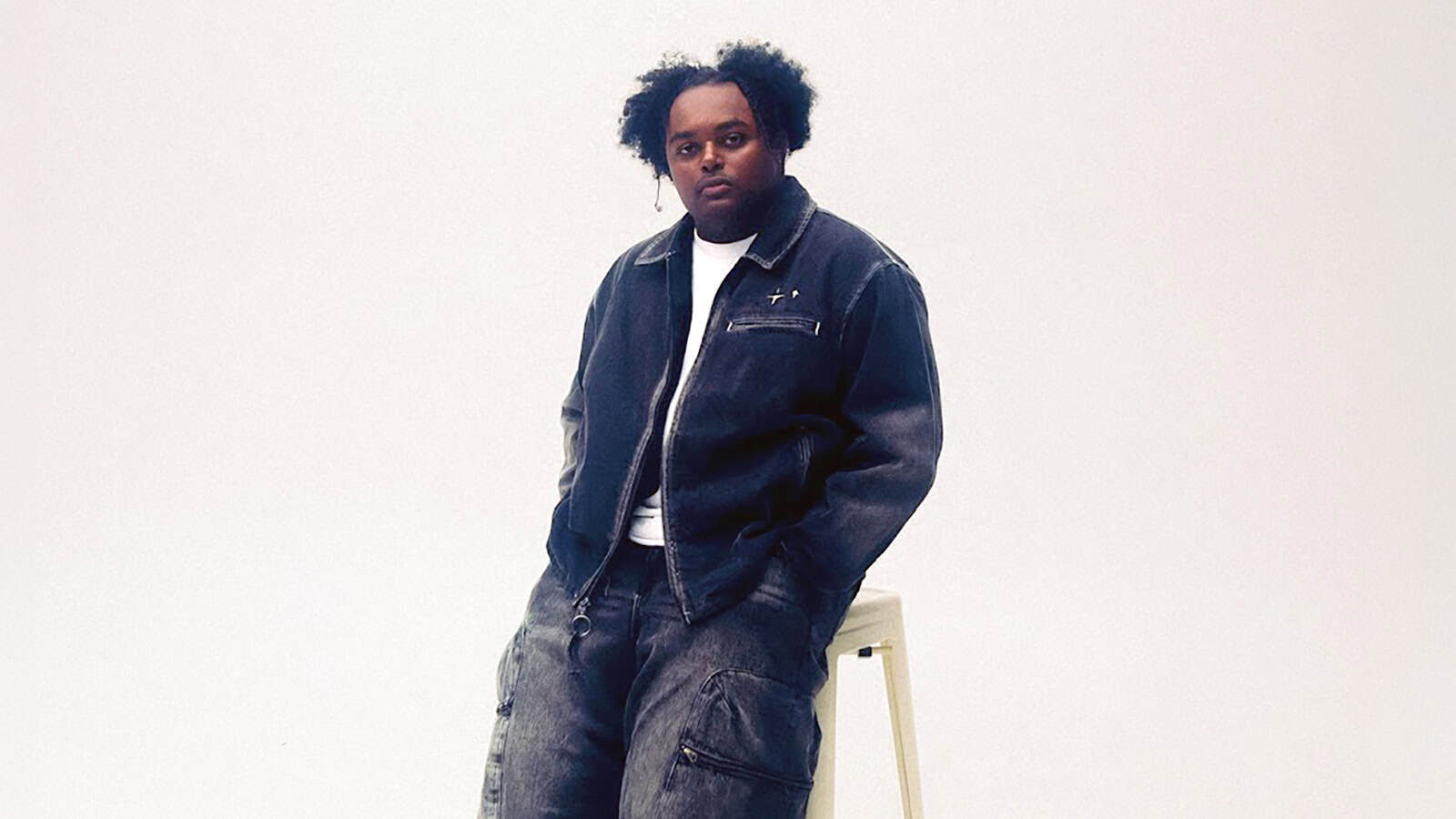 A kid in a dark denim jacket and cargo jeans, channeling hip-hop vibes, sits on a white stool against a plain light background.