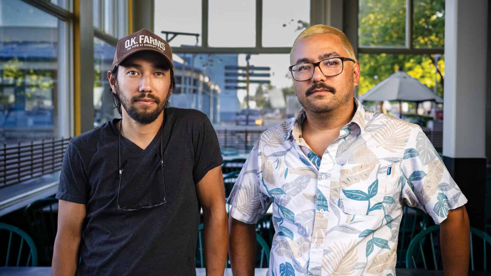 Two men, Seth Pacleb and Zach Pacleb of System Smashers, stand side by side in a casual indoor setting with large windows; one wears a hat and dark shirt, the other has a patterned shirt and glasses.
