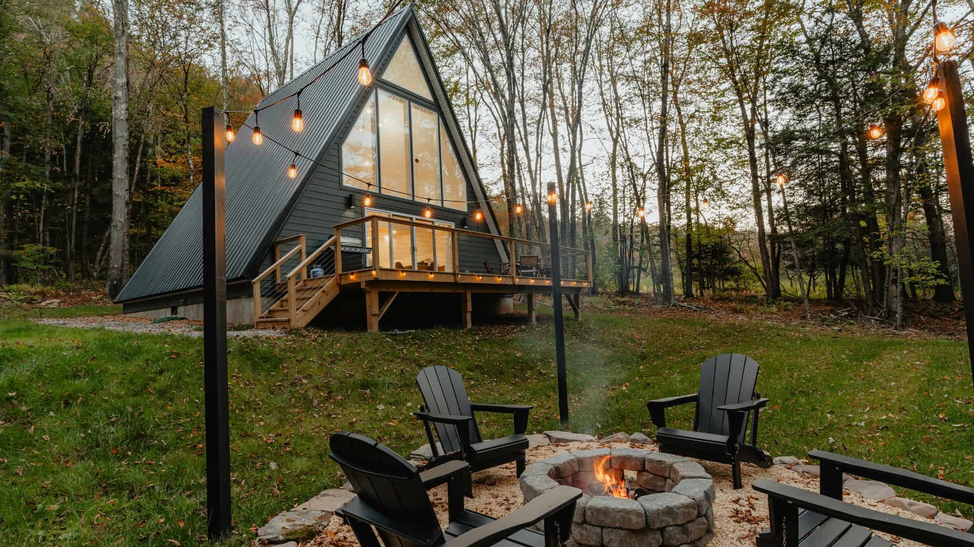 A-frame cabin with large windows and a deck surrounded by trees, featuring a fire pit area with four black chairs and string lights.