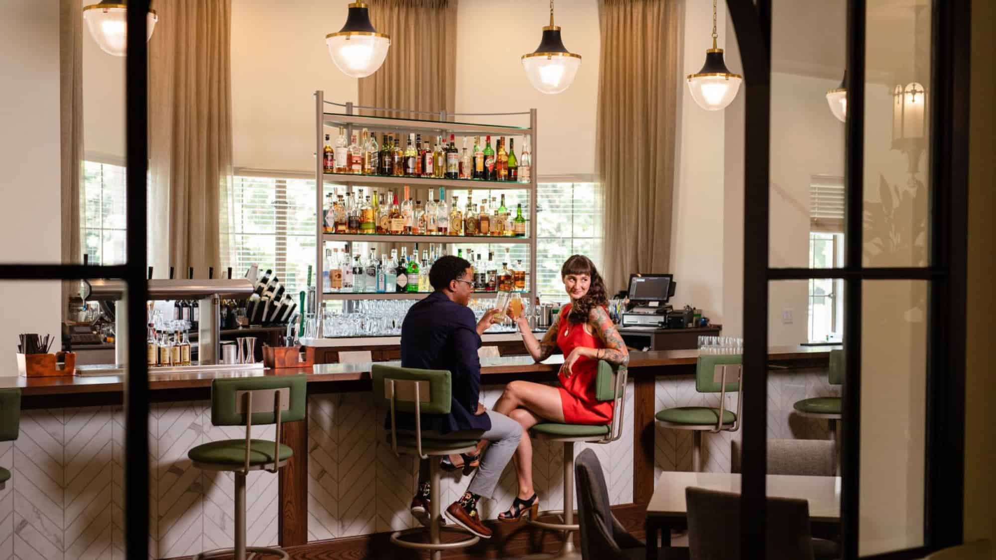 Two people sit at a bar, facing each other and holding drinks, with bottles and glasses displayed on shelves behind the counter.
