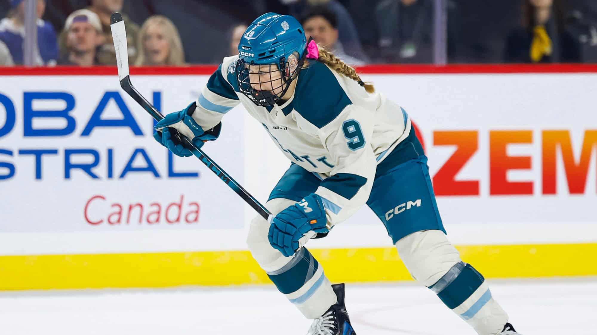 Danielle Serdachny, wearing white and teal gear with the number 9, skates on the ice in a Women's Hockey League game, holding her hockey stick as spectators watch from the background.