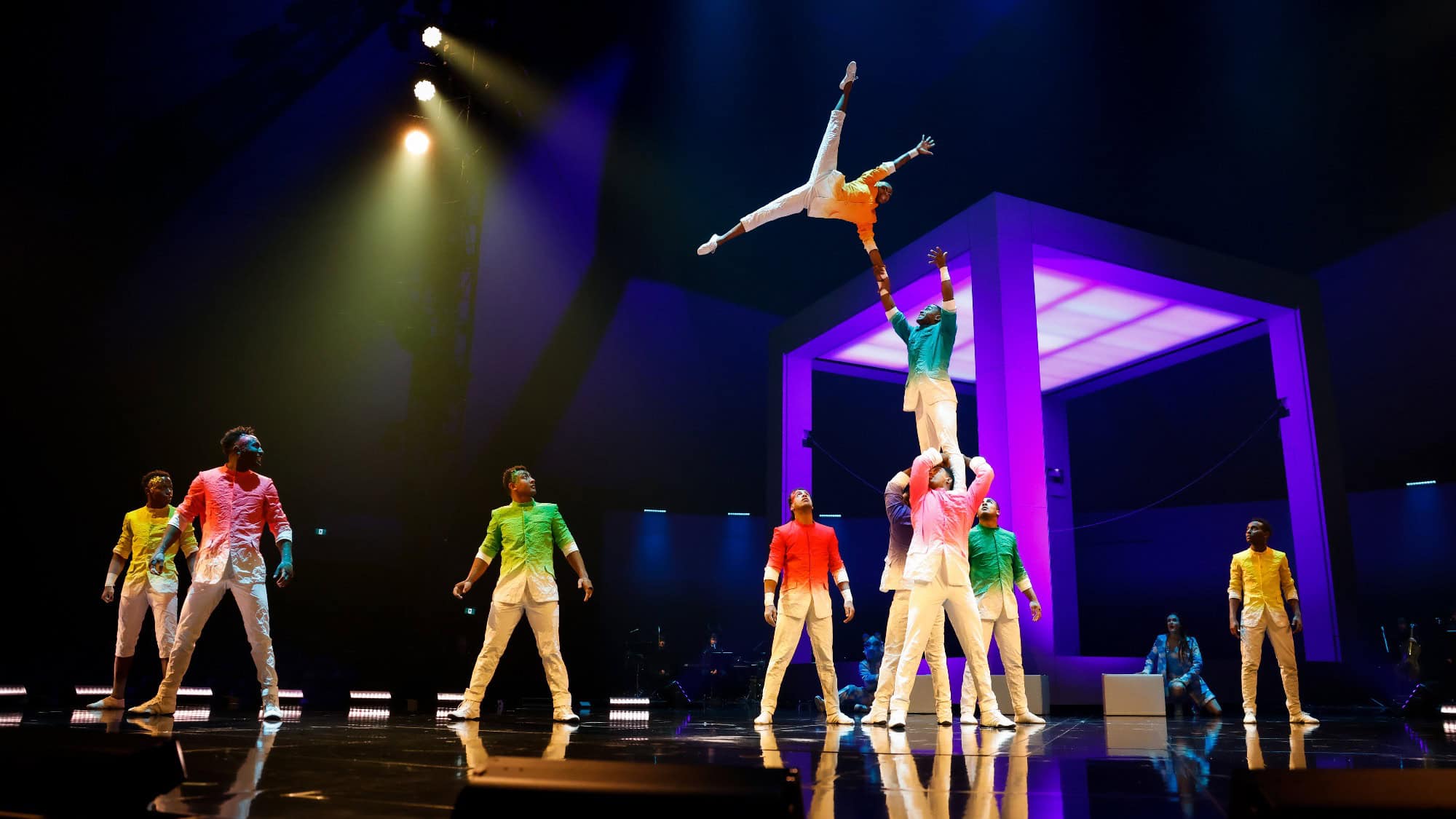 A group of acrobats in colorful costumes perform a balancing act on stage under dramatic lighting, with one performer lifted high above the others.