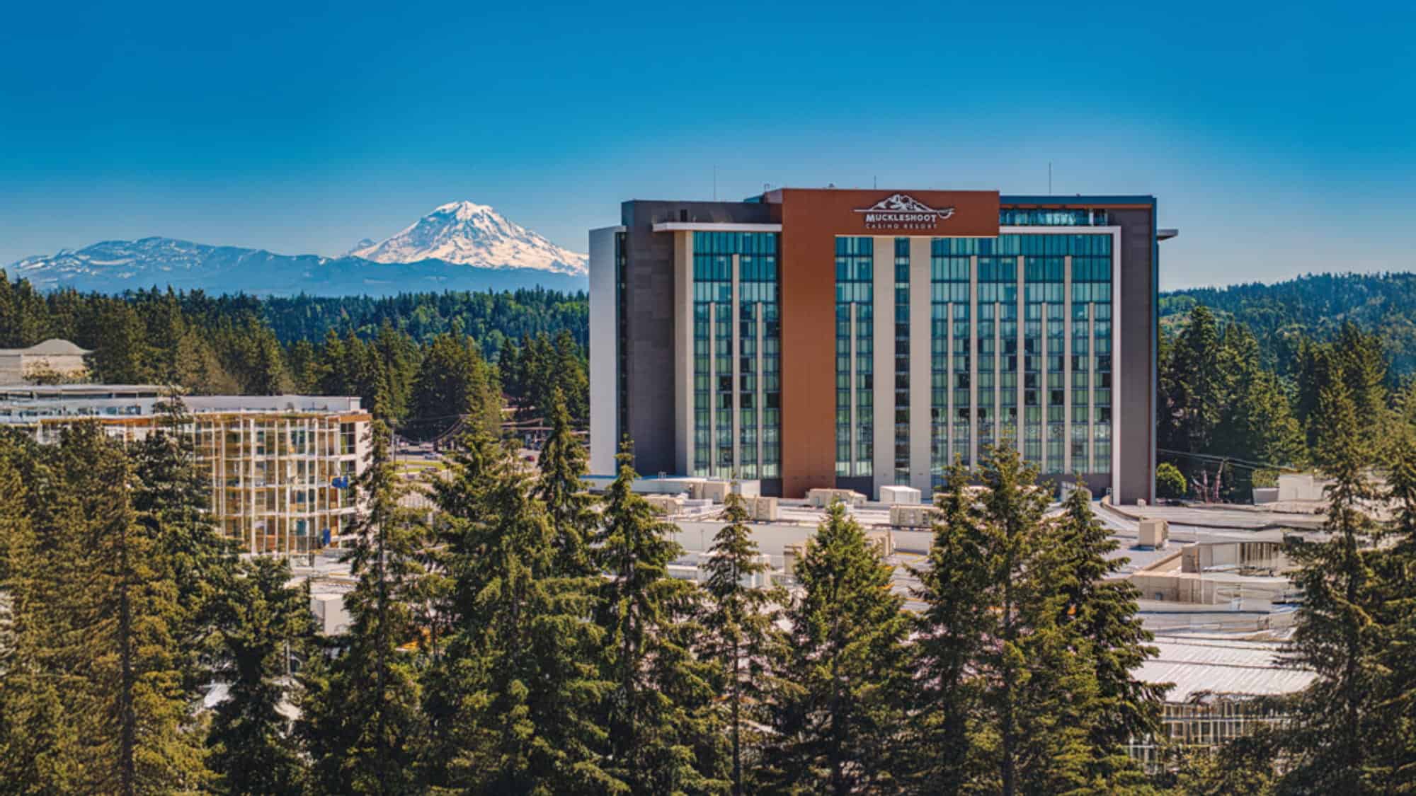A modern multi-story building with a mountain logo and “MultiCare” on top, surrounded by trees, stands proudly with a snow-capped mountain in the background under a clear blue sky—truly care Like Nowhere Else.