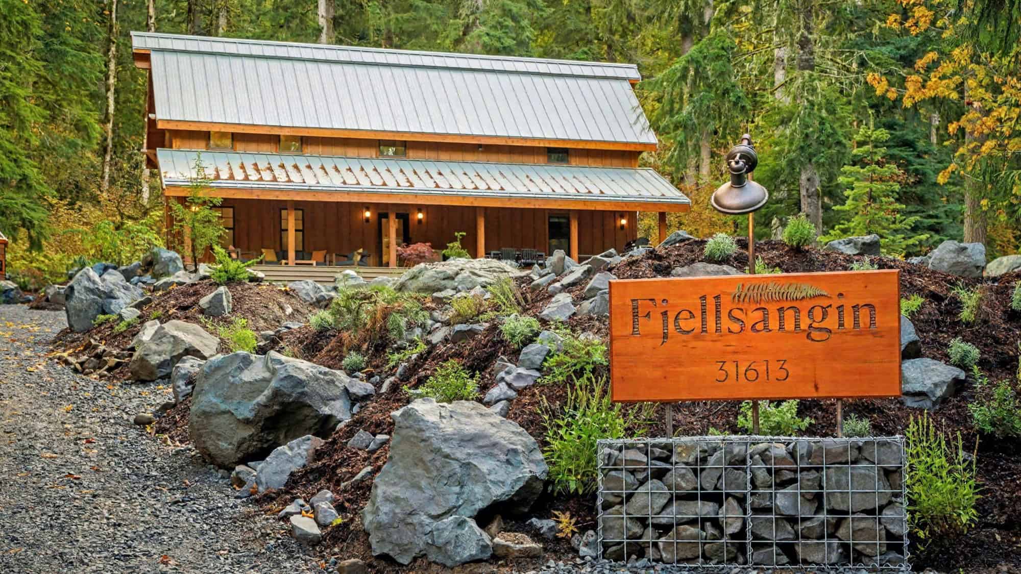 A wooden house with a metal roof is built into the trees in a forested area, with a large sign reading "Fjellsangin 31613" in the foreground among rocks and shrubs.