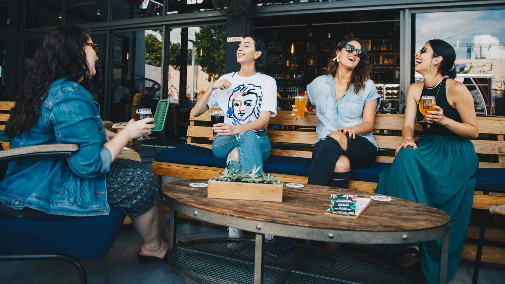 Four women sit on outdoor benches at a bar, holding drinks and laughing together around a table with a small plant centerpiece.