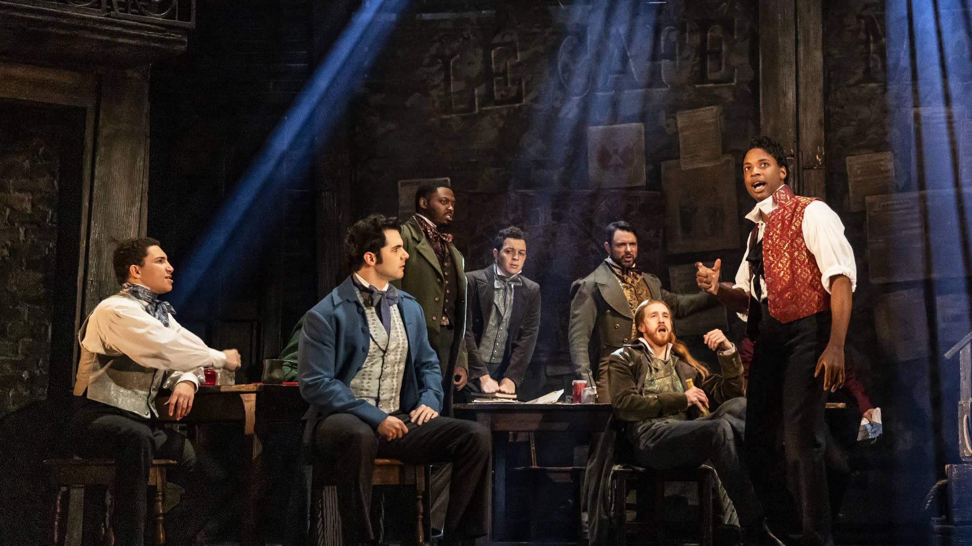 A group of men in period costumes sit and stand around a table on a dimly lit stage, with one man in a red vest standing and speaking.