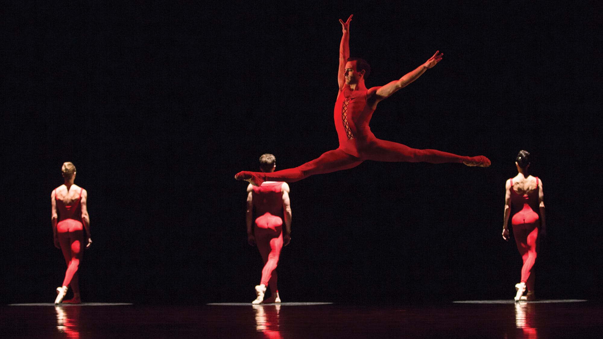 Four ballet dancers in red outfits perform on stage; one leaps mid-air with arms and legs extended, while three others face away toward the background—a striking moment from the vibrant arts in Seattle.