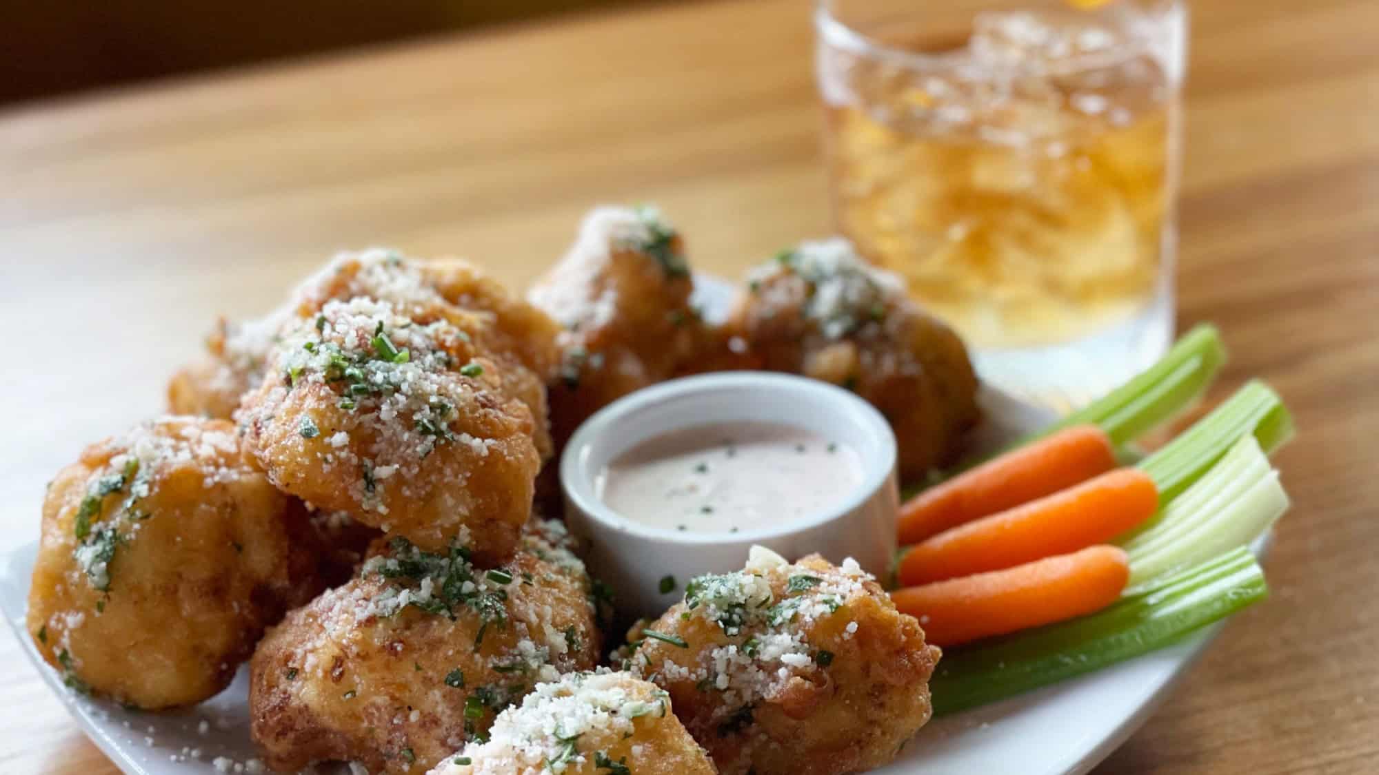A plate of breaded, fried cauliflower bites with a side of dipping sauce, celery sticks, and carrot sticks, next to a glass of iced beverage on a wooden table.