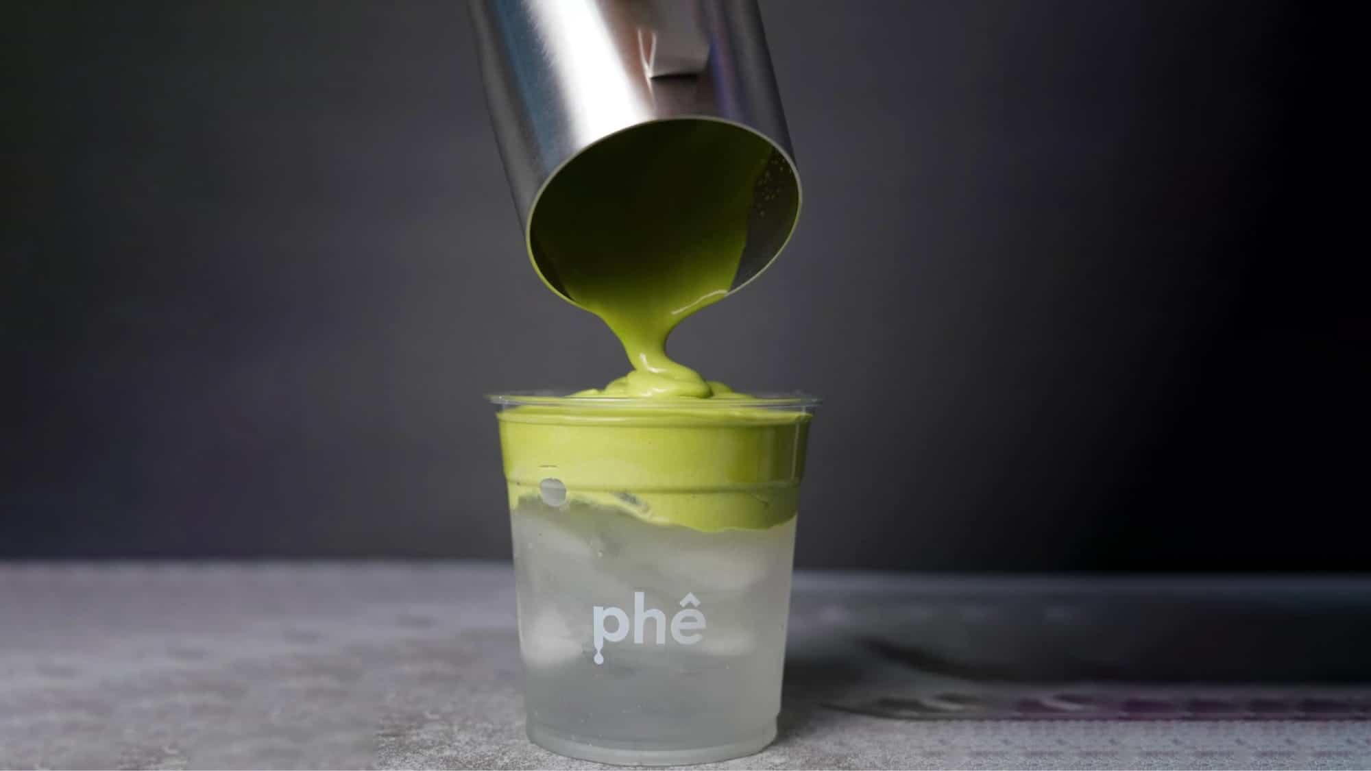 A metal cup pours thick green matcha cream over ice in a clear plastic cup labeled "phê," showcasing a unique twist on Vietnamese coffee against a dark background—perfect for fans of Seattle coffee shops seeking something new.