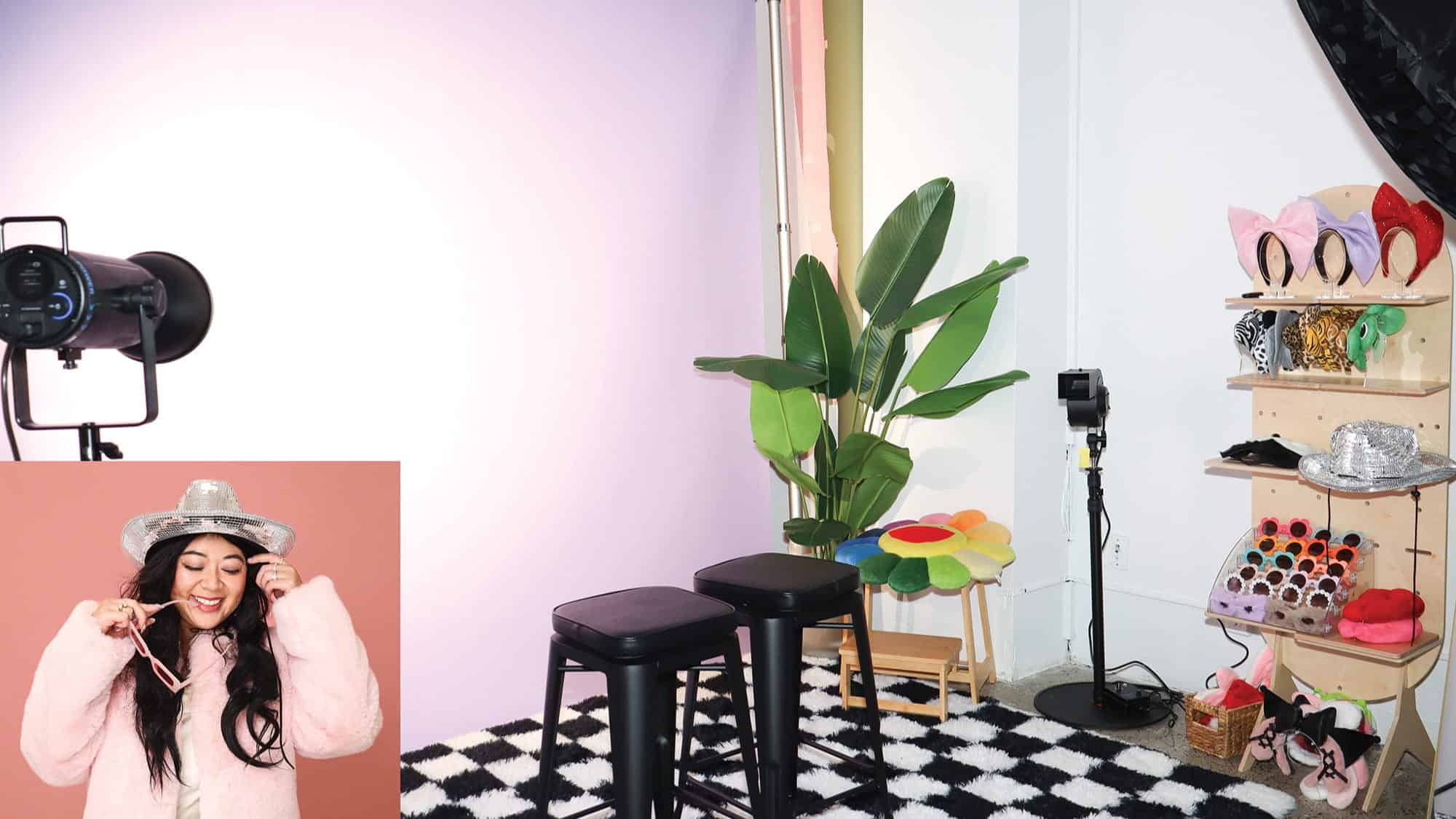 A photo studio setup with stools, lighting equipment, a plant, and accessories on shelves offers space to play; inset shows a woman with long dark hair smiling and holding a necklace, wearing a silver hat.