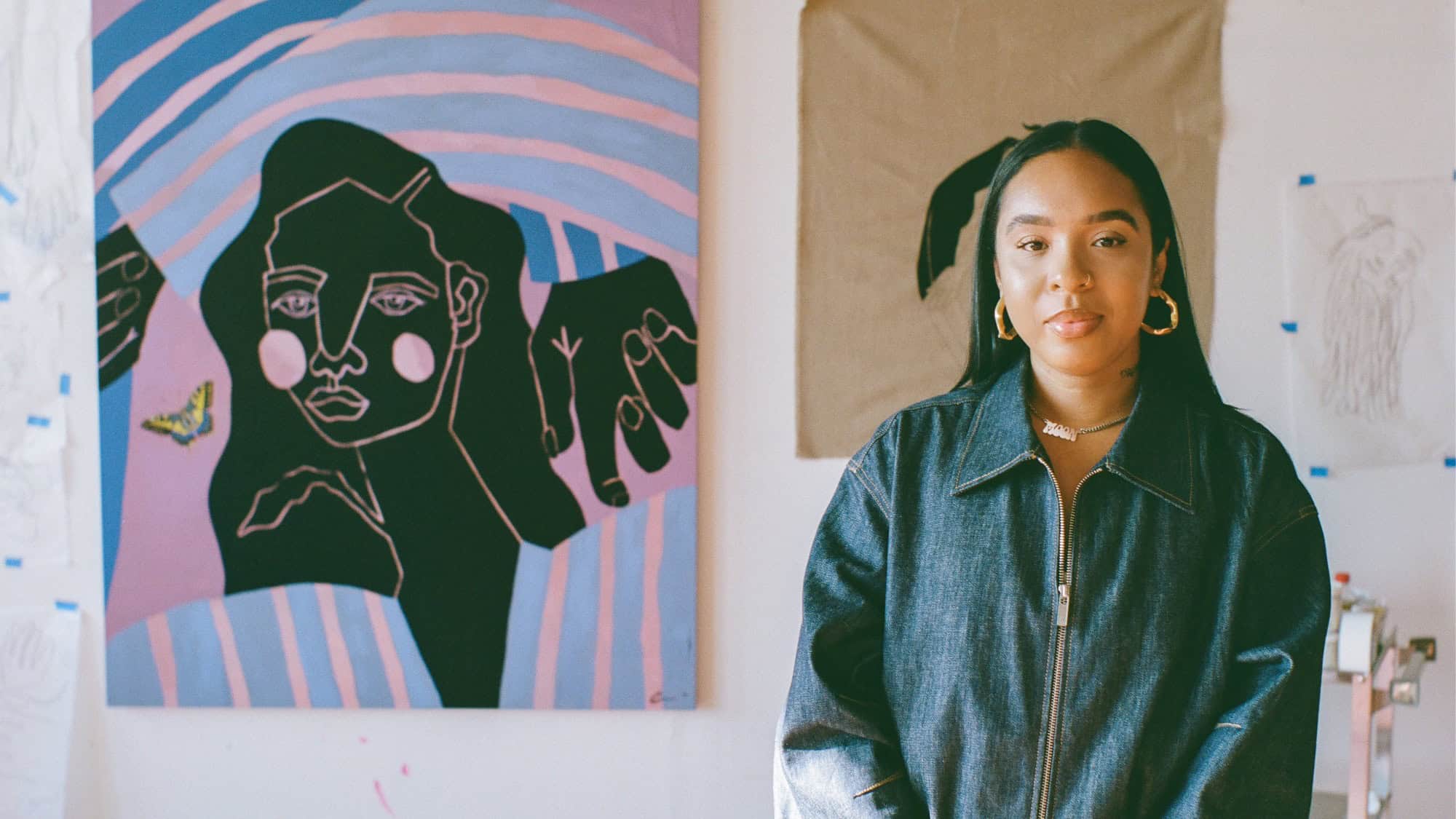 A woman in a denim jacket stands in front of colorful abstract artwork and sketches displayed on a wall.