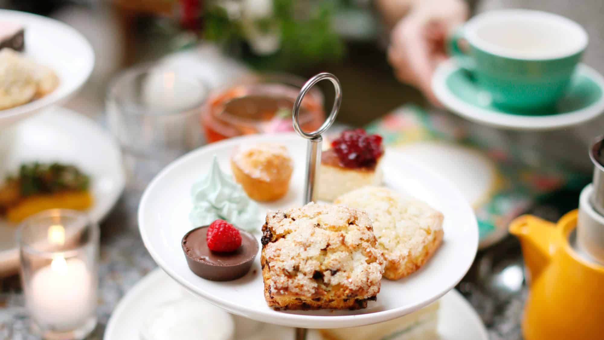 A tiered tray with assorted pastries, scones, jam, and a chocolate dessert; a hand holds a green teacup in the background.
