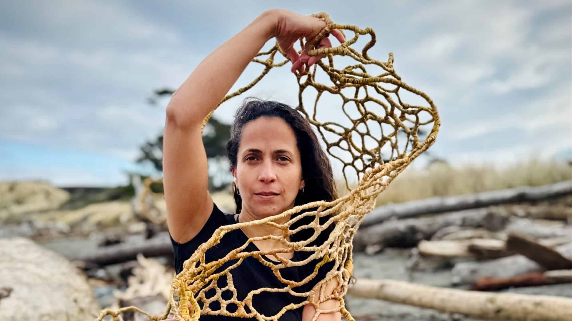 A person holds up an intricate, net-like woven structure outdoors, with driftwood and cloudy sky in the background.