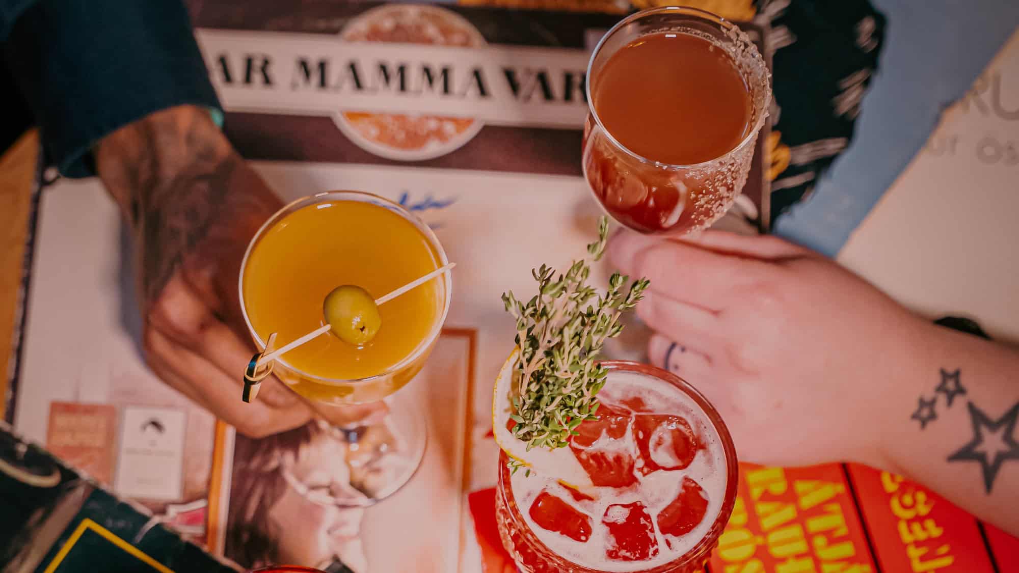 Three hands holding different colorful cocktails over a table with menus and magazines. One drink has an olive garnish, another has herbs, and a third is served in a coupe glass.