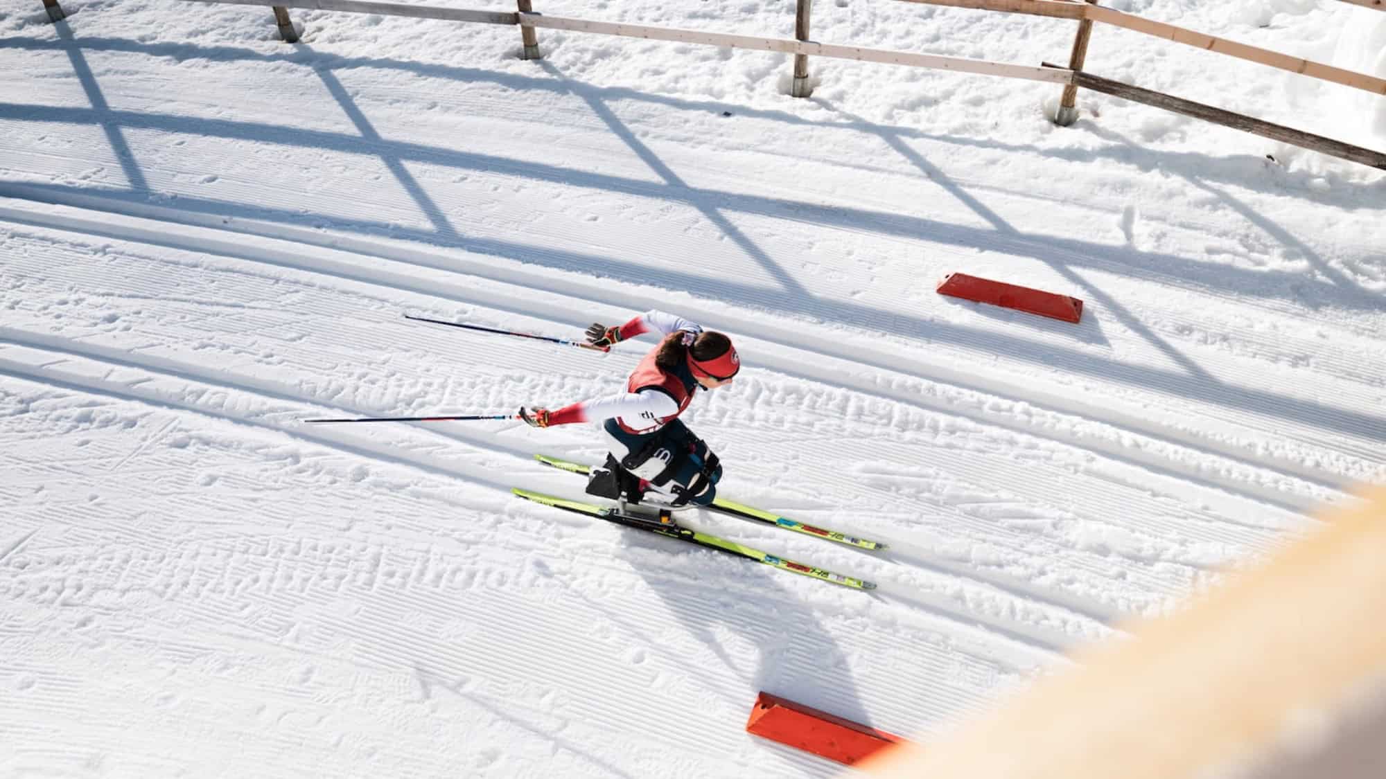 A skier wearing a red and white outfit glides along groomed cross-country ski tracks on a snowy course bordered by a wooden fence.