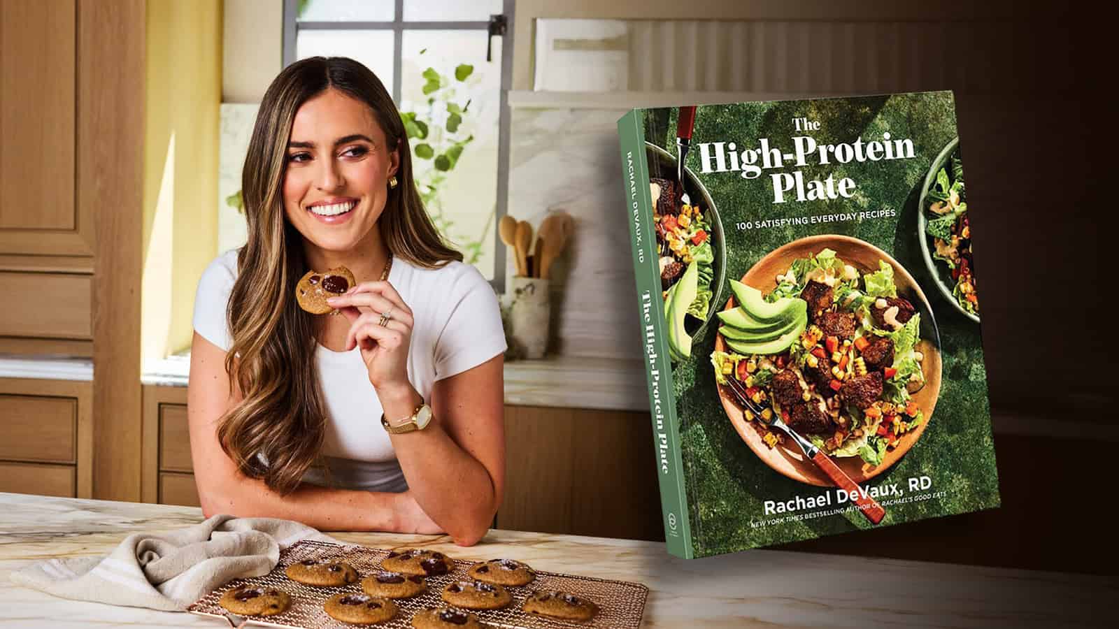 A woman sits at a kitchen counter eating a cookie, with a plate of cookies in front of her. Next to her is the cookbook "The High-Protein Plate" by Rachael DeVaux, RD.