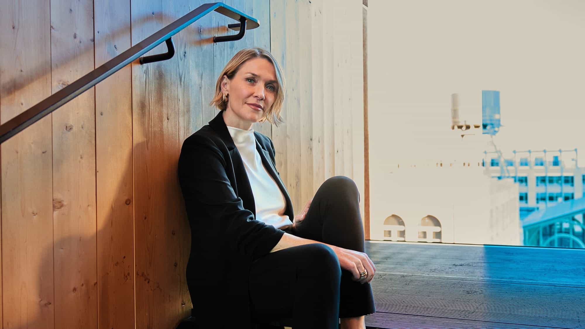 A woman in a black suit and white top sits on stairs next to a wooden wall, highlighting the building connection and modern design, with an urban view visible in the background.