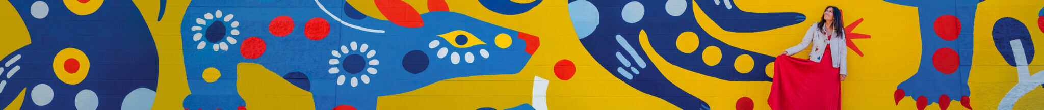 A woman in a red and white outfit stands against a colorful mural featuring abstract blue and red shapes with floral patterns on a bright yellow background.