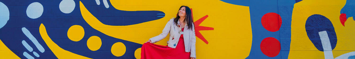 A woman in a beige jacket and red skirt stands smiling in front of a colorful mural with abstract blue, yellow, and red shapes.