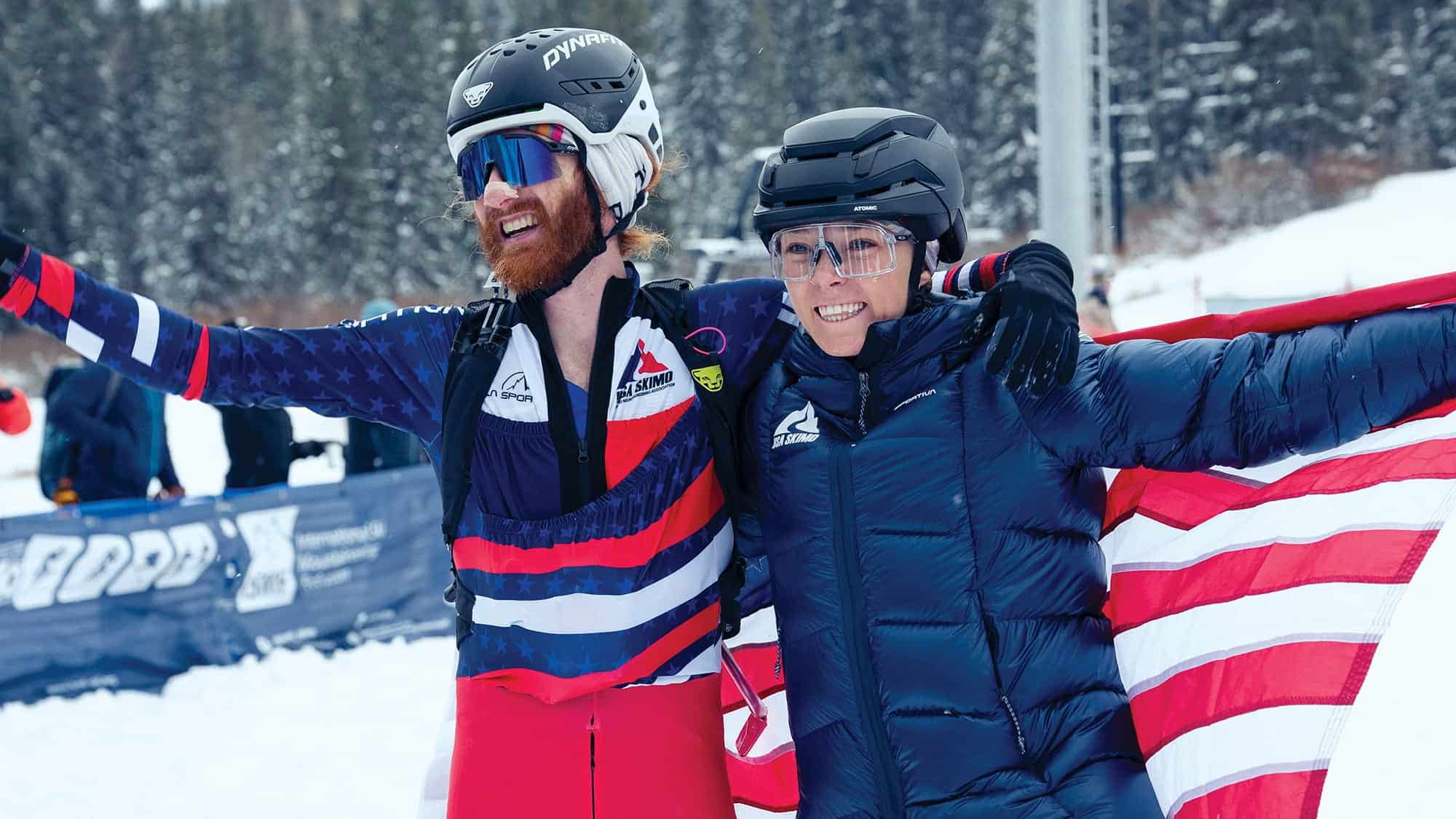 Two athletes in winter gear, including skimo professional runner Anna Gibson in a racing suit, stand arm in arm and smile on a snowy slope, proudly holding a U.S. flag.