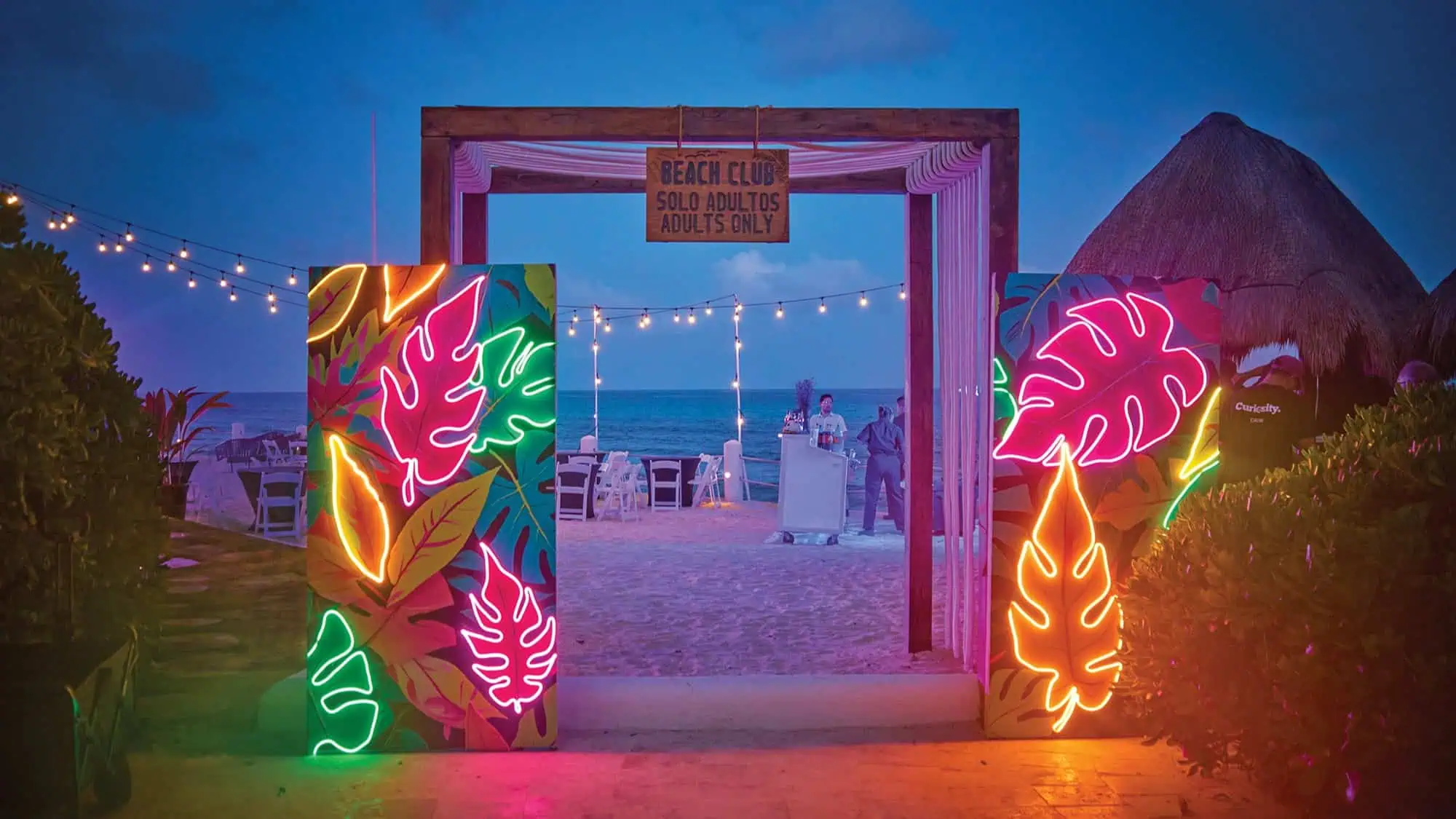 A neon-lit entrance with tropical leaf designs leads to an adults-only beach club at dusk, where string lights and the ocean evoke the relaxation and wellness in Mexico found at top all-inclusive resorts.
