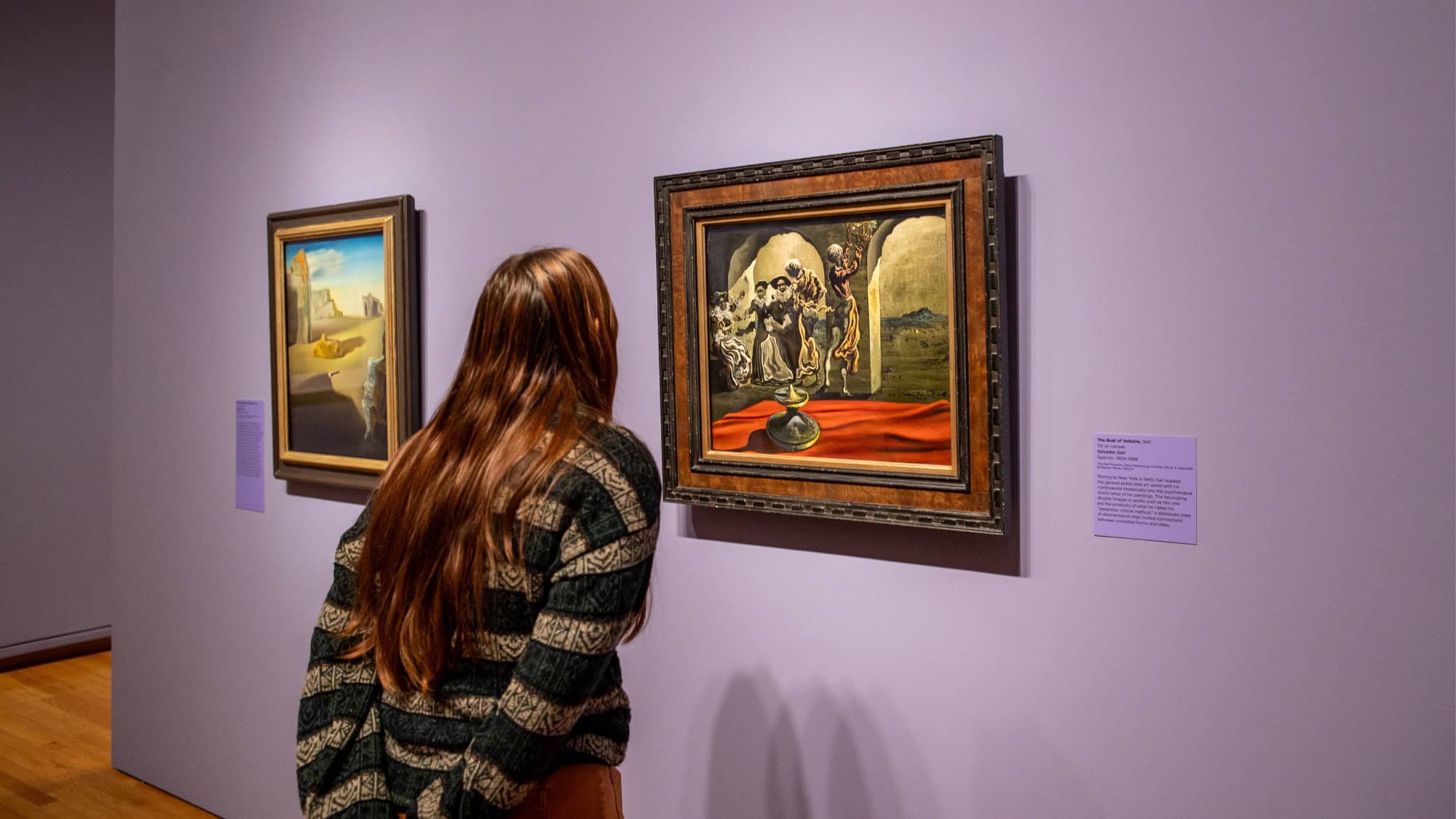A person with long brown hair closely observes a framed painting on display in an art gallery with purple walls.