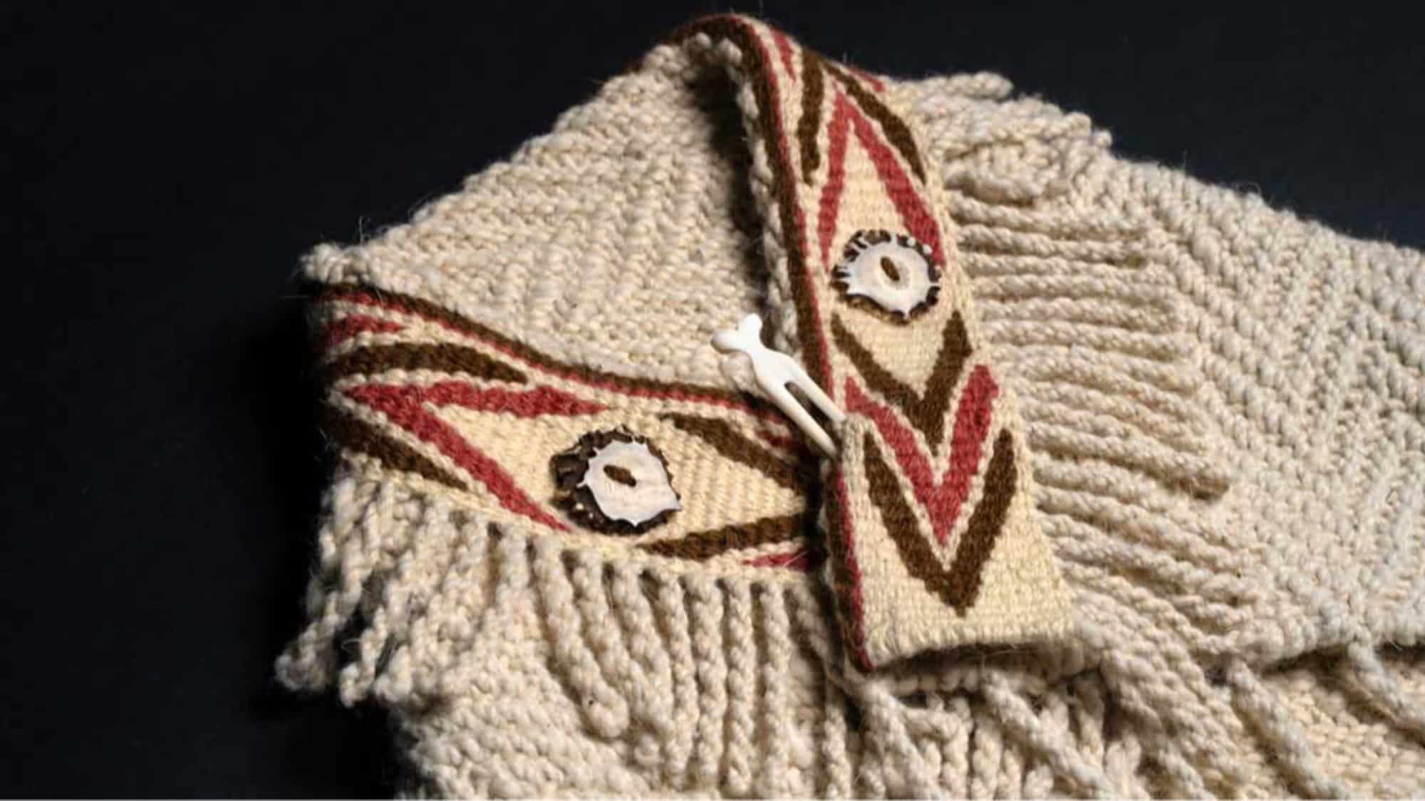 A close-up of a knitted cream-colored garment with decorative patterned trim, fringes, and a button-and-loop closure.